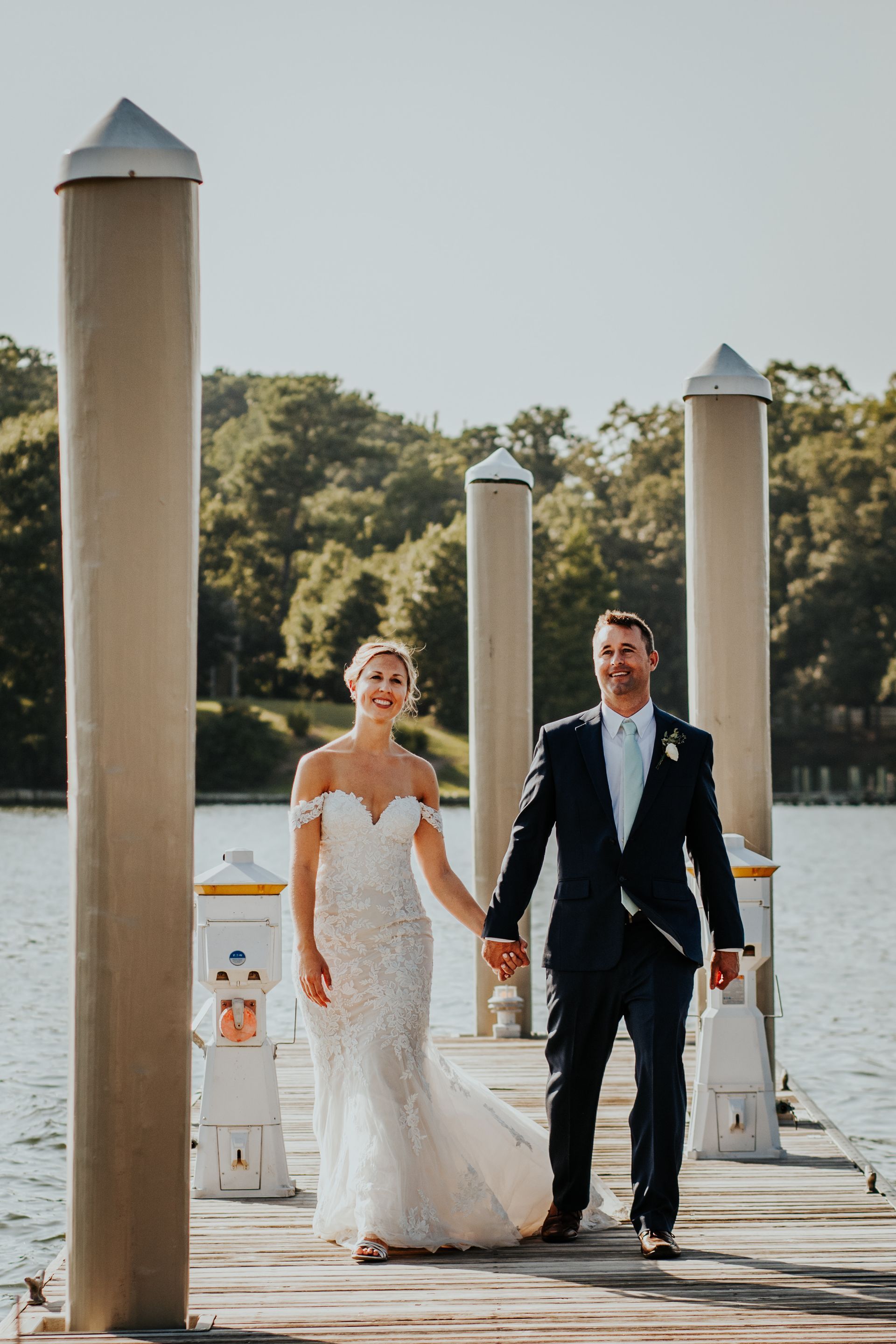 A bride and groom are walking on a dock holding hands.