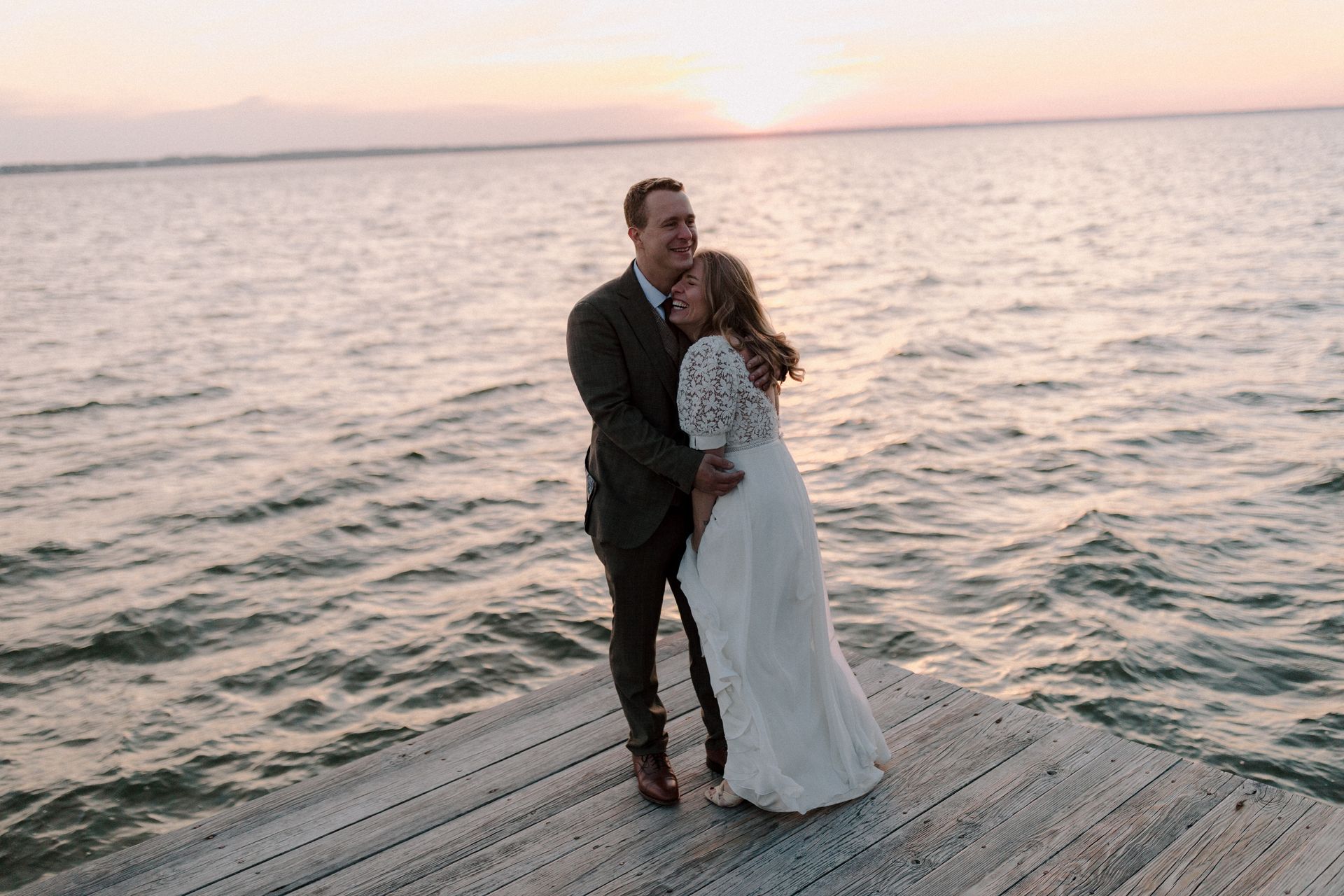 A bride and groom are standing on a dock near the ocean at sunset.