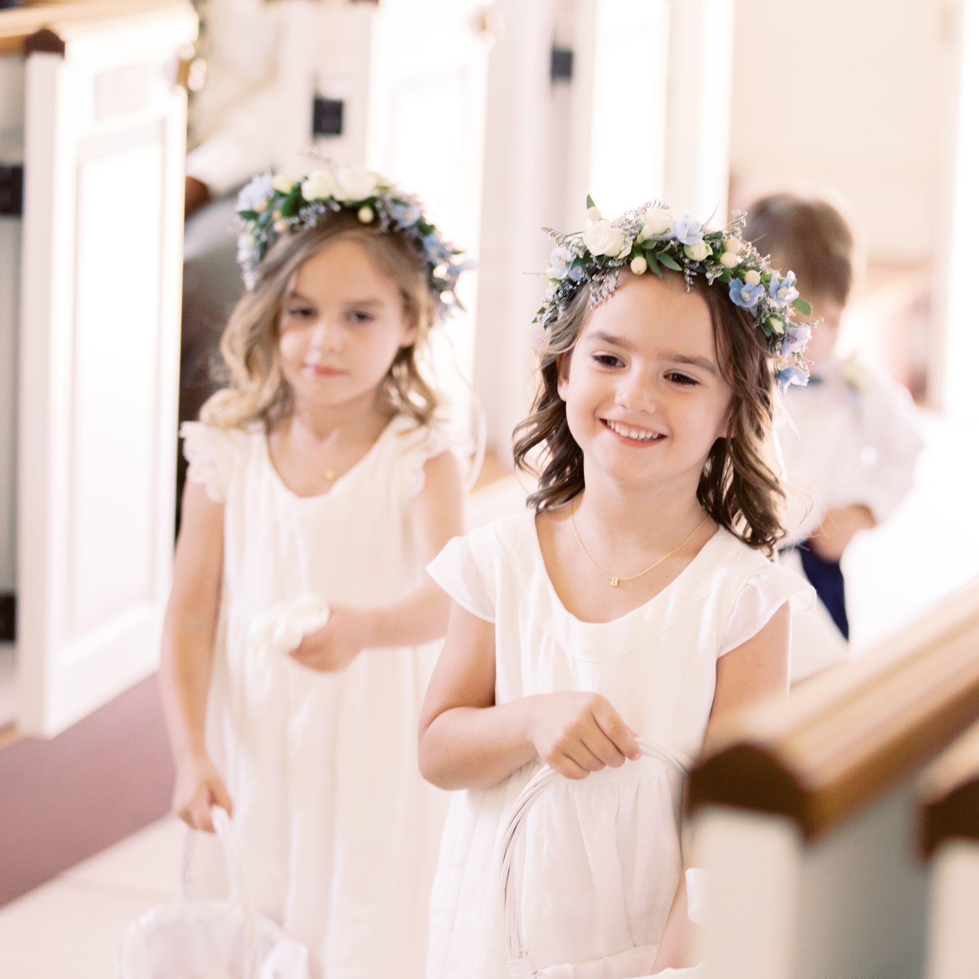Two flower girls wearing flower crowns are walking down a hallway.