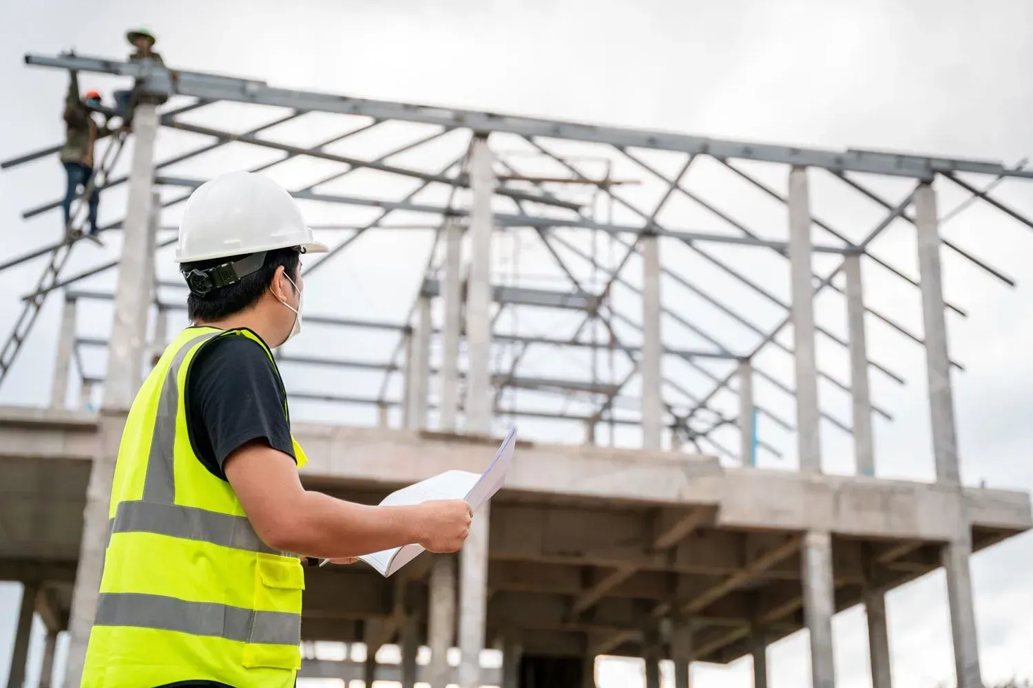 Construction worker in white hard hat and yellow vest inspecting a steel frame building under construction