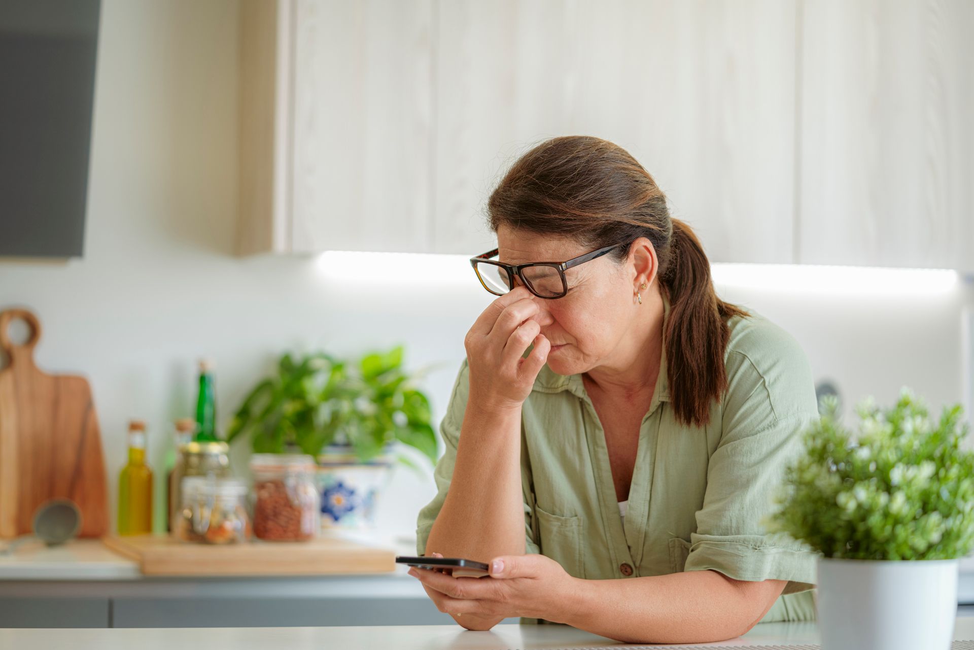 Woman in glasses holding phone, rubbing her nose with a pained expression in a kitchen.