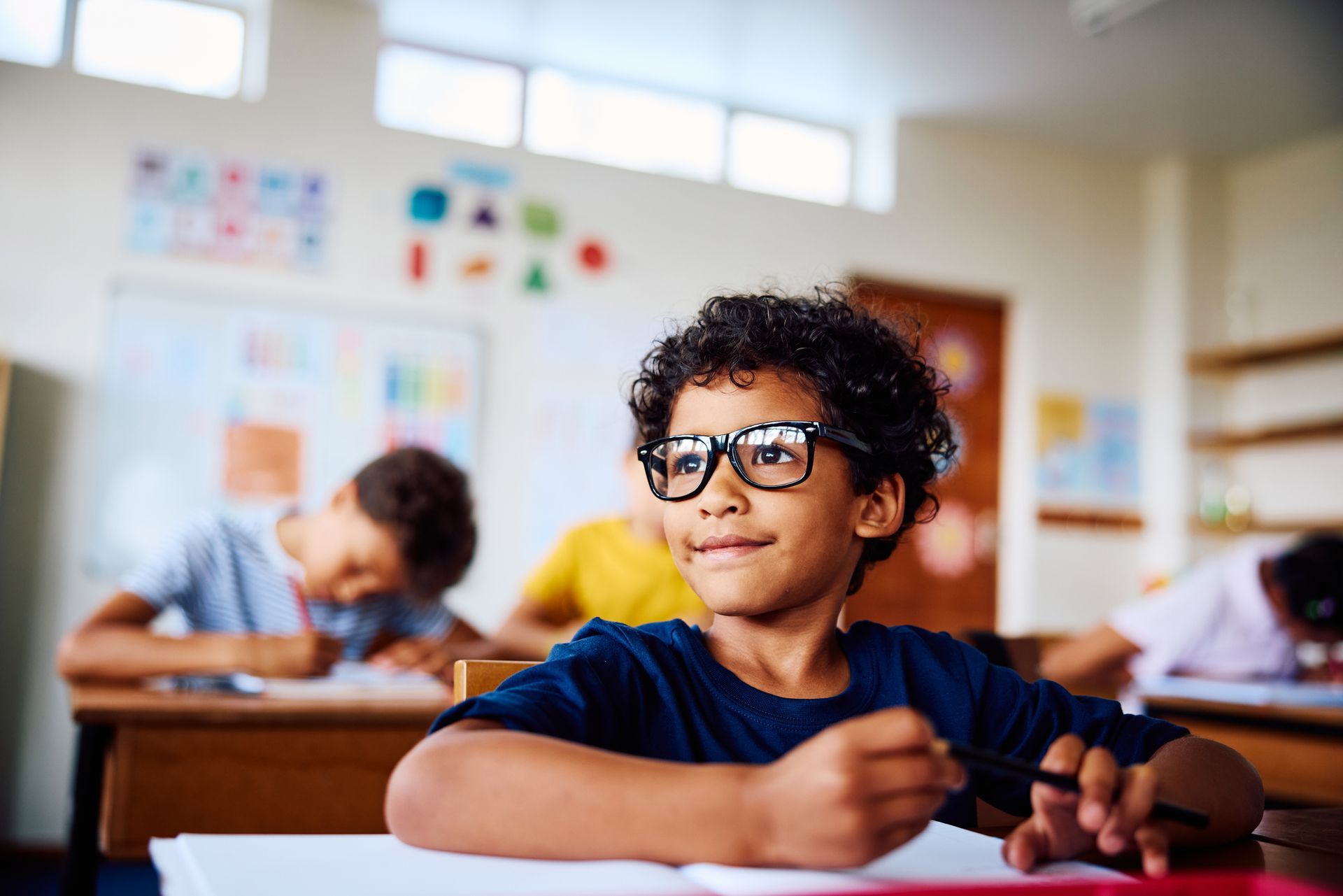 Boy with glasses smiles in a classroom, holding a pencil, looking upward. Other students are in the background.