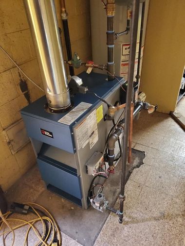 Blue furnace with metal vent pipes in a basement utility area with unfinished walls and exposed pipes