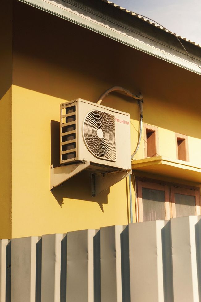 Wall-mounted air conditioner on a yellow house exterior above a gray fence.
