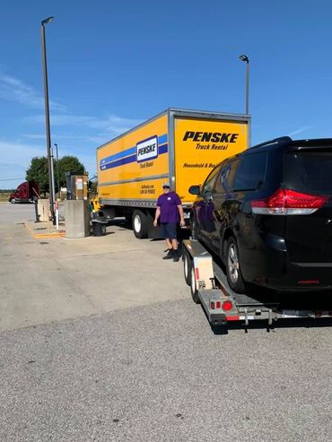 Penske moving truck and black car on a trailer at a gas station. Man in purple shirt walks towards truck.