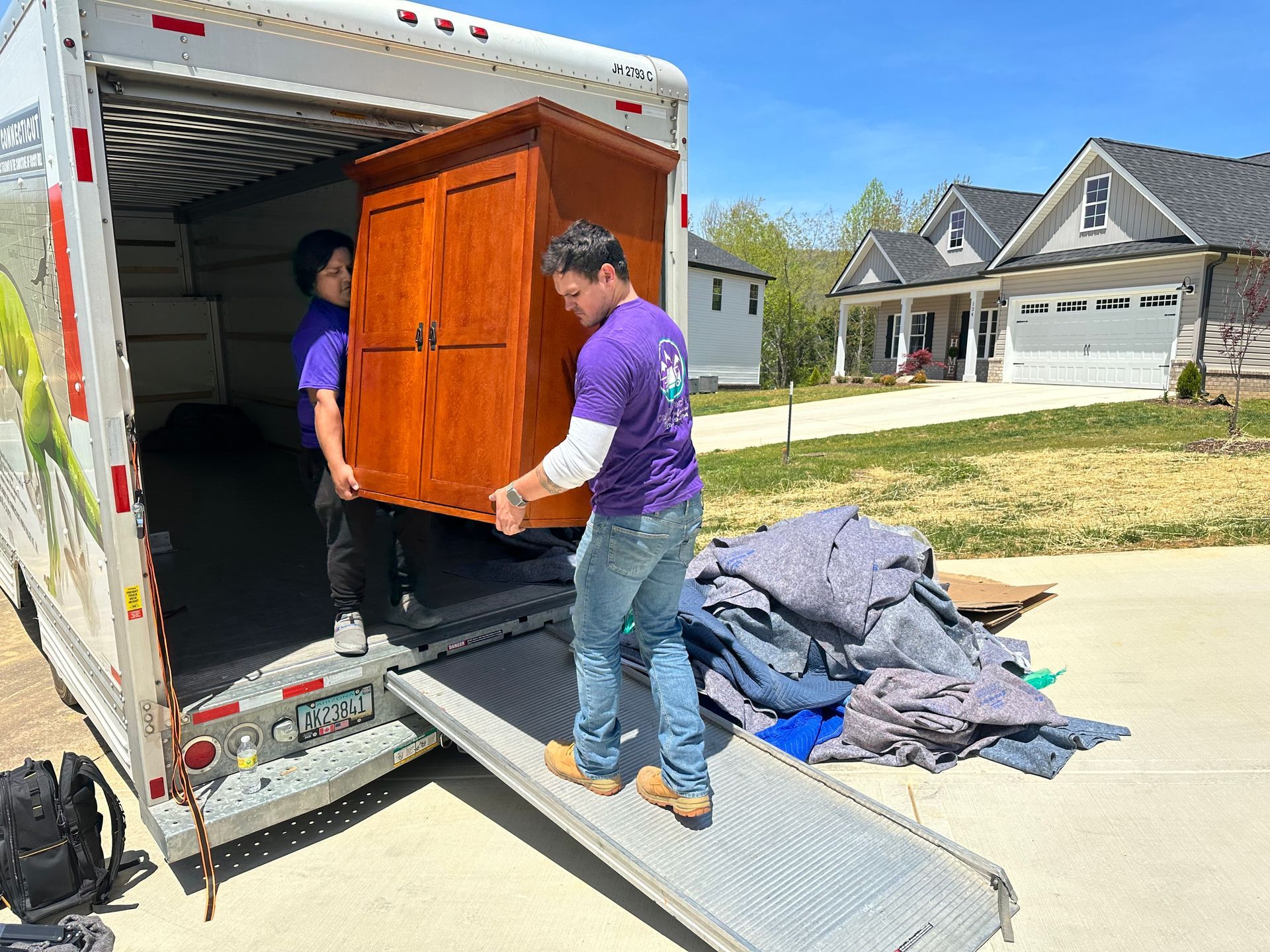 Two movers unload a wooden cabinet from a truck onto a ramp in a suburban driveway.