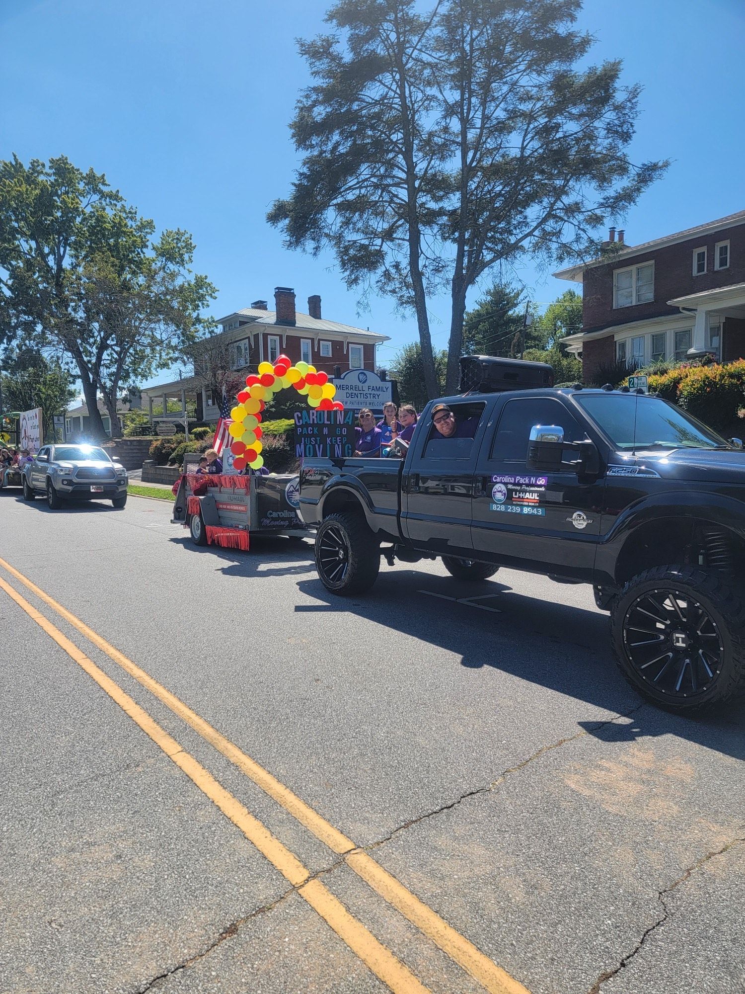 Black truck towing a trailer with a red, yellow, and orange balloon arch in a parade on a sunny day.