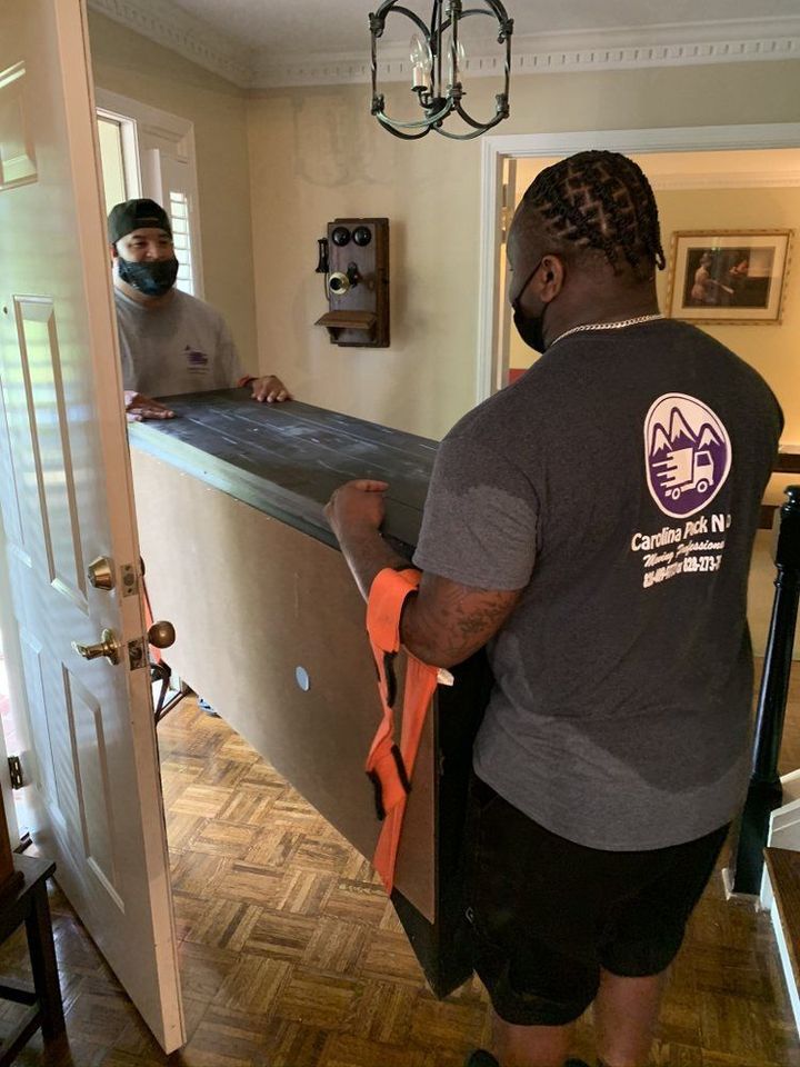 Two movers wearing masks carry a large, heavy piece of furniture through a doorway.