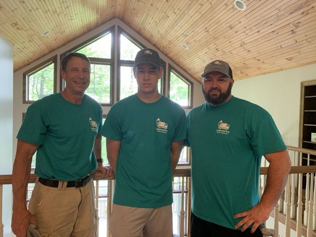 Three men in teal shirts and caps stand inside a home.