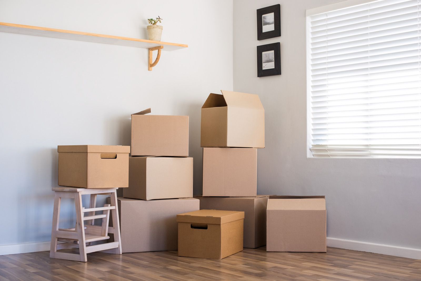 Cardboard moving boxes stacked in a room, near a small shelf and a window with blinds.
