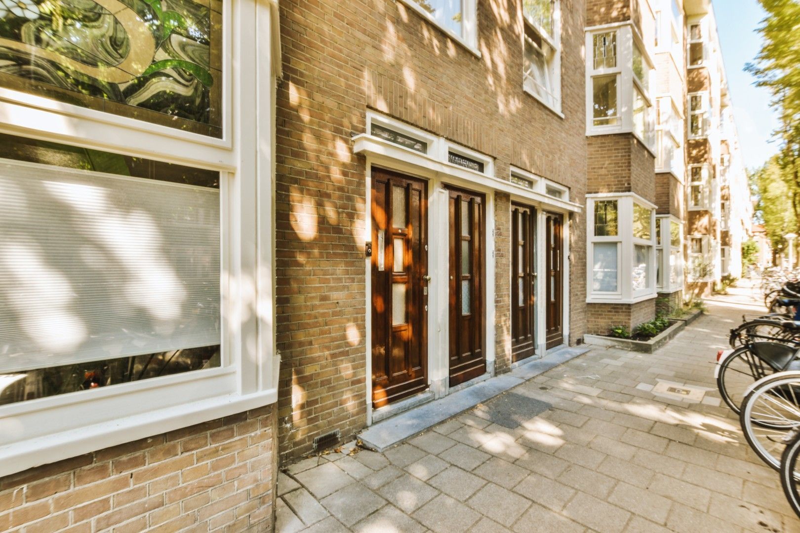Smalle bakstenen ingang van het appartementencomplex met glazen deuren, een zonnig trottoir en fietsen die buiten geparkeerd staan.