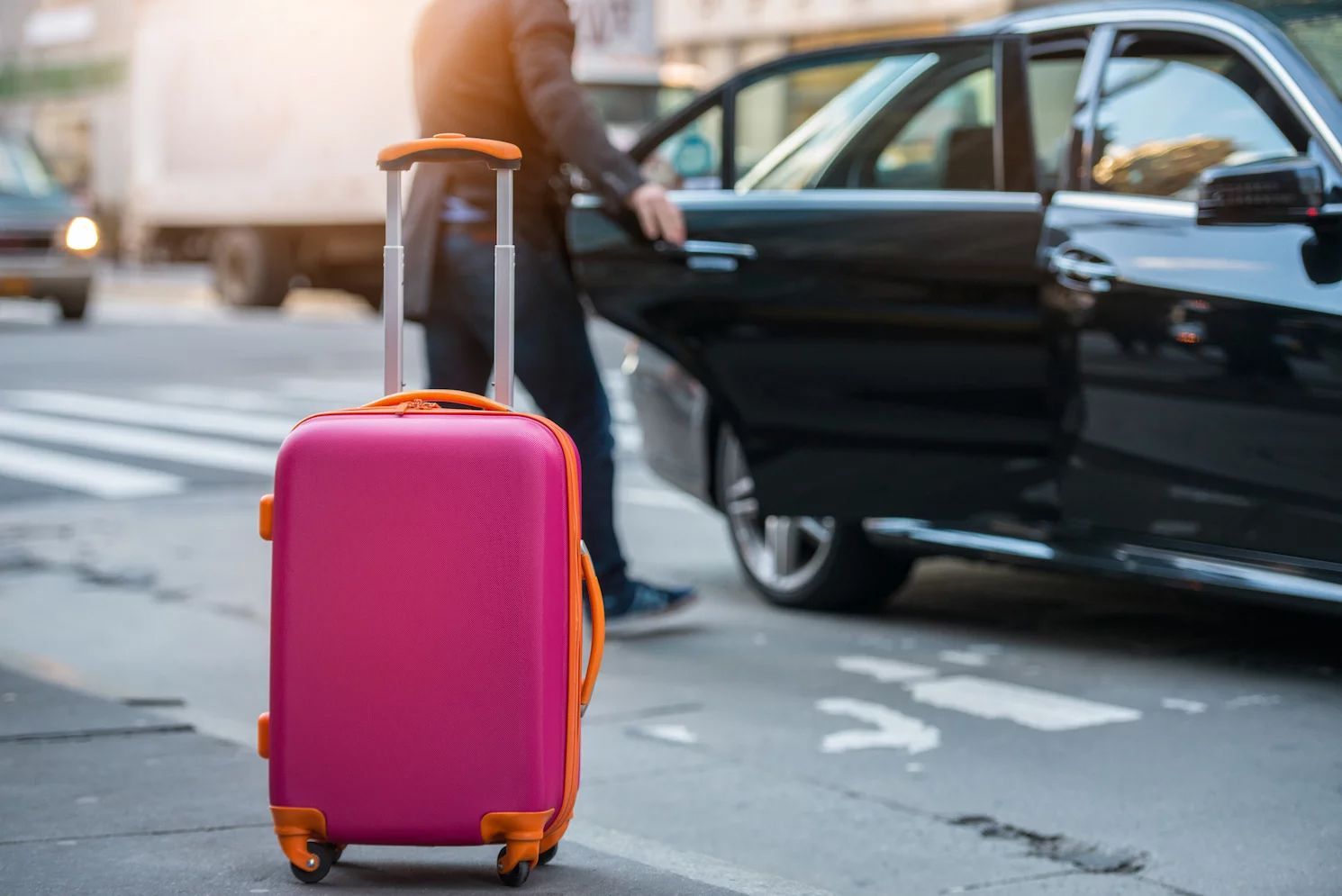 Pink suitcase beside a person opening a black car door on a city street