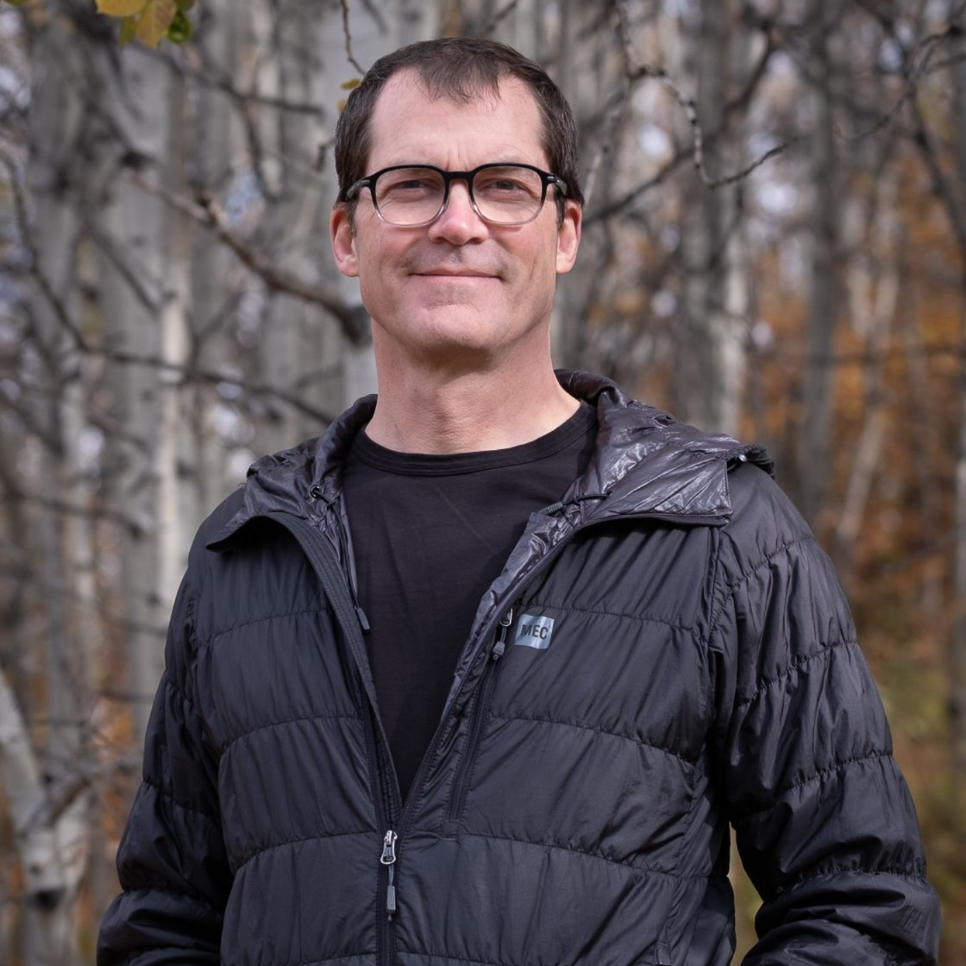 Man wearing glasses and black jacket stands in front of fall foliage.