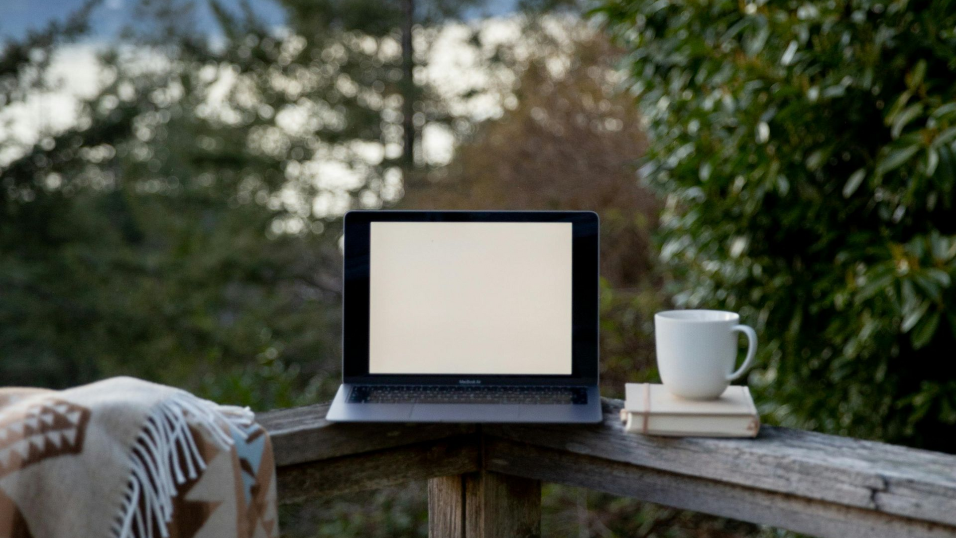 Laptop with blank screen, coffee mug, and books on outdoor railing overlooking greenery.