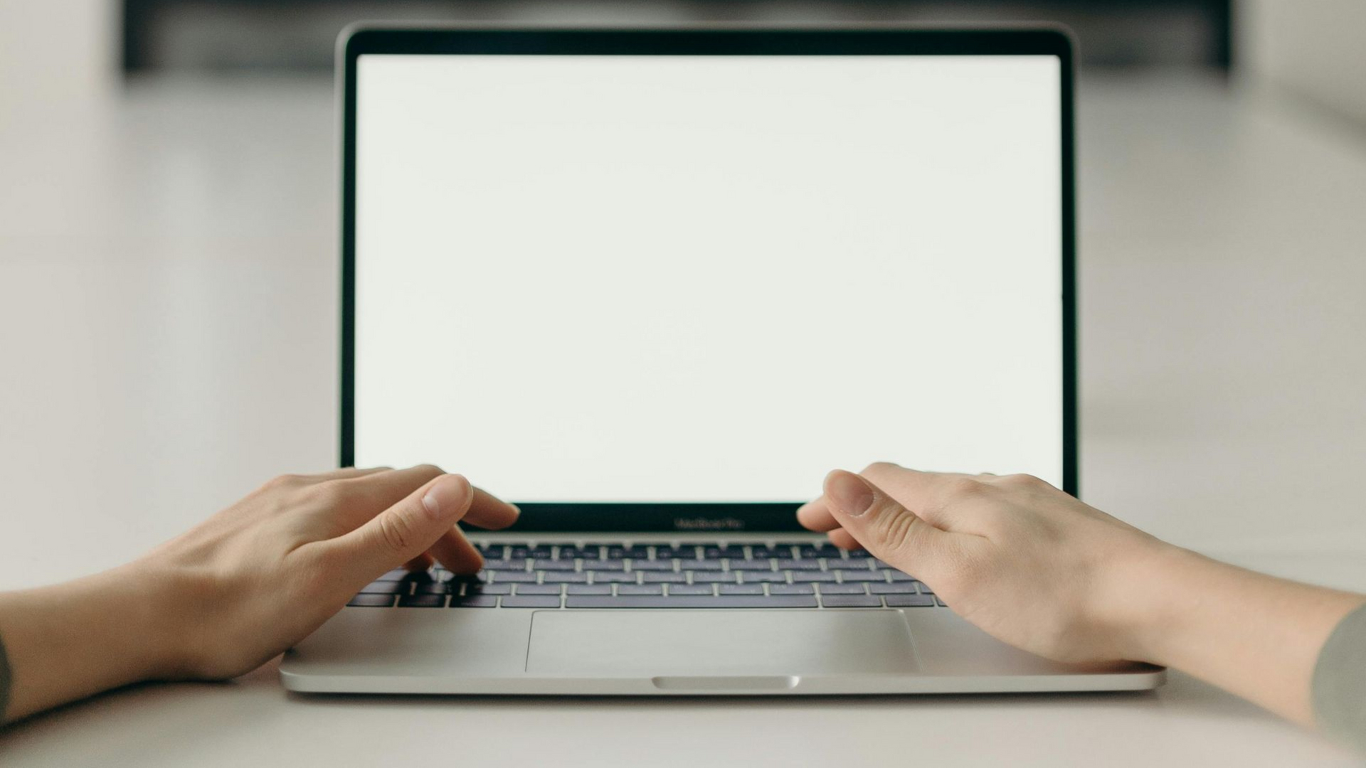 Hands typing on a laptop with a blank white screen, sitting on a white surface.