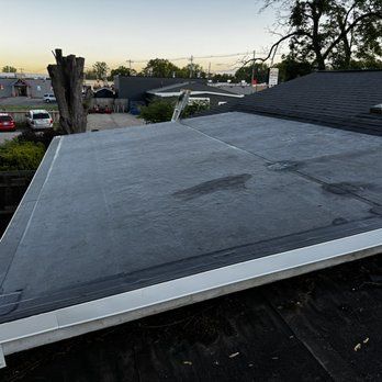 Flat, dark gray roof with white trim; view from an adjacent sloped roof; overcast sky in background.
