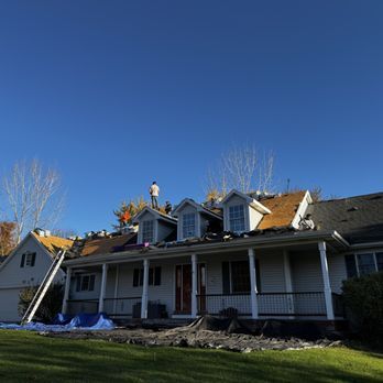 Roofers working on a residential roof, sunny day, blue tarp below, ladder on the side.