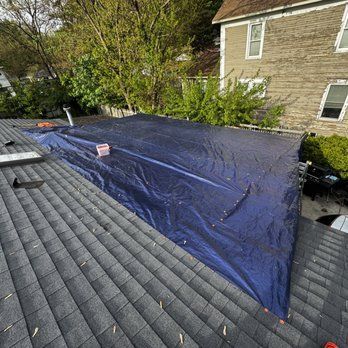 Blue tarp covering a roof with asphalt shingles; a building and trees are in the background.