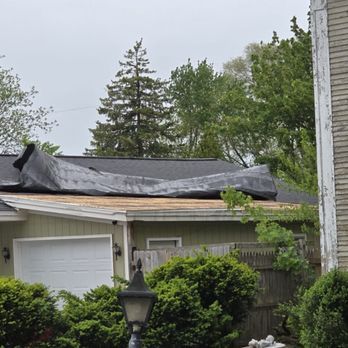 A damaged roof covered with black tarp. A white garage door and green shrubs are in the foreground.