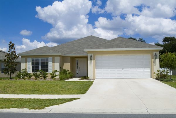 Beige house with a two-car garage, white garage door, and a green lawn under a blue sky with clouds.