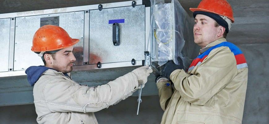 Two men wearing hard hats are working on a ventilation system.