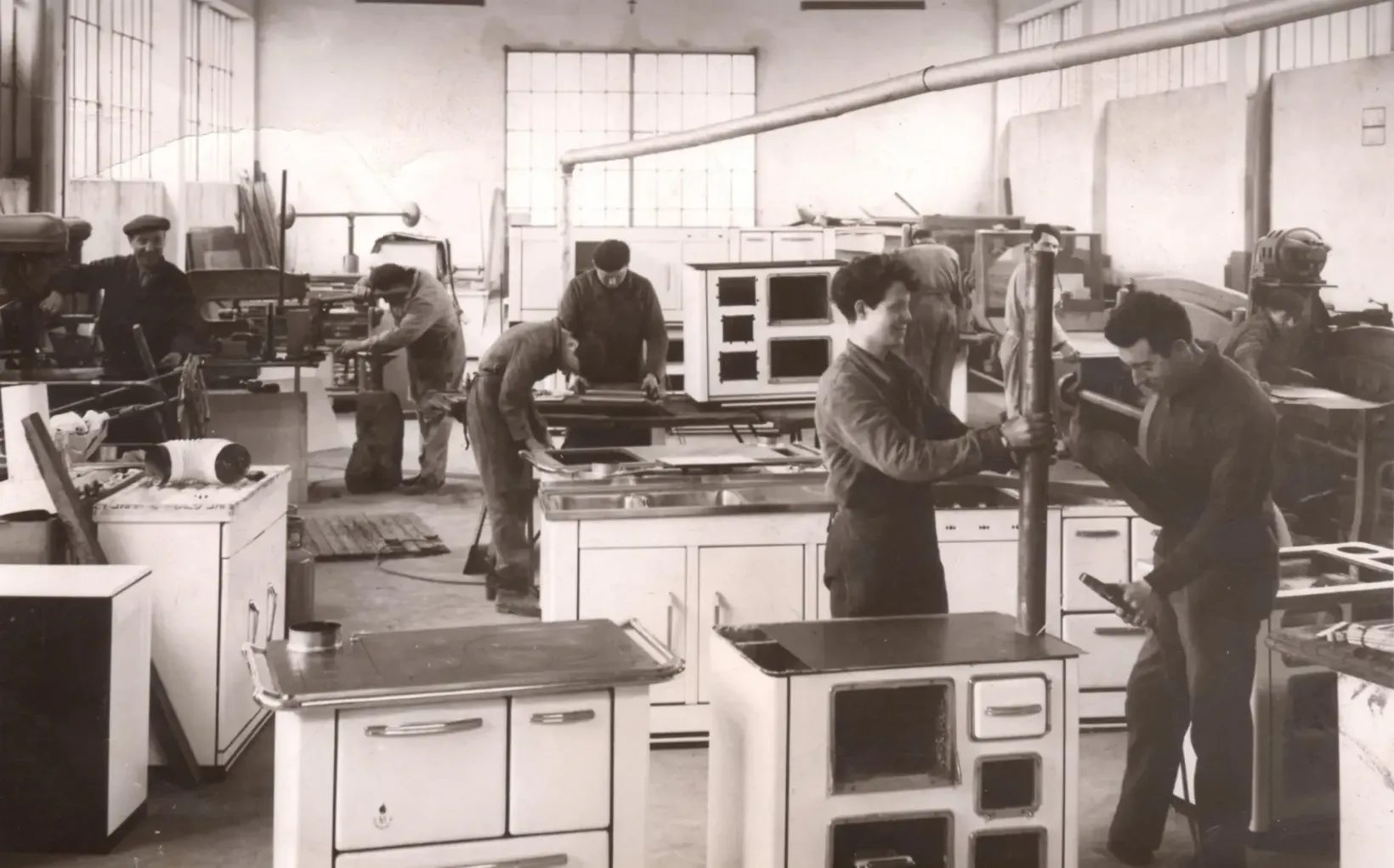 Officina con diversi uomini che costruiscono stufe. Lavori di carpenteria e assemblaggio in corso.