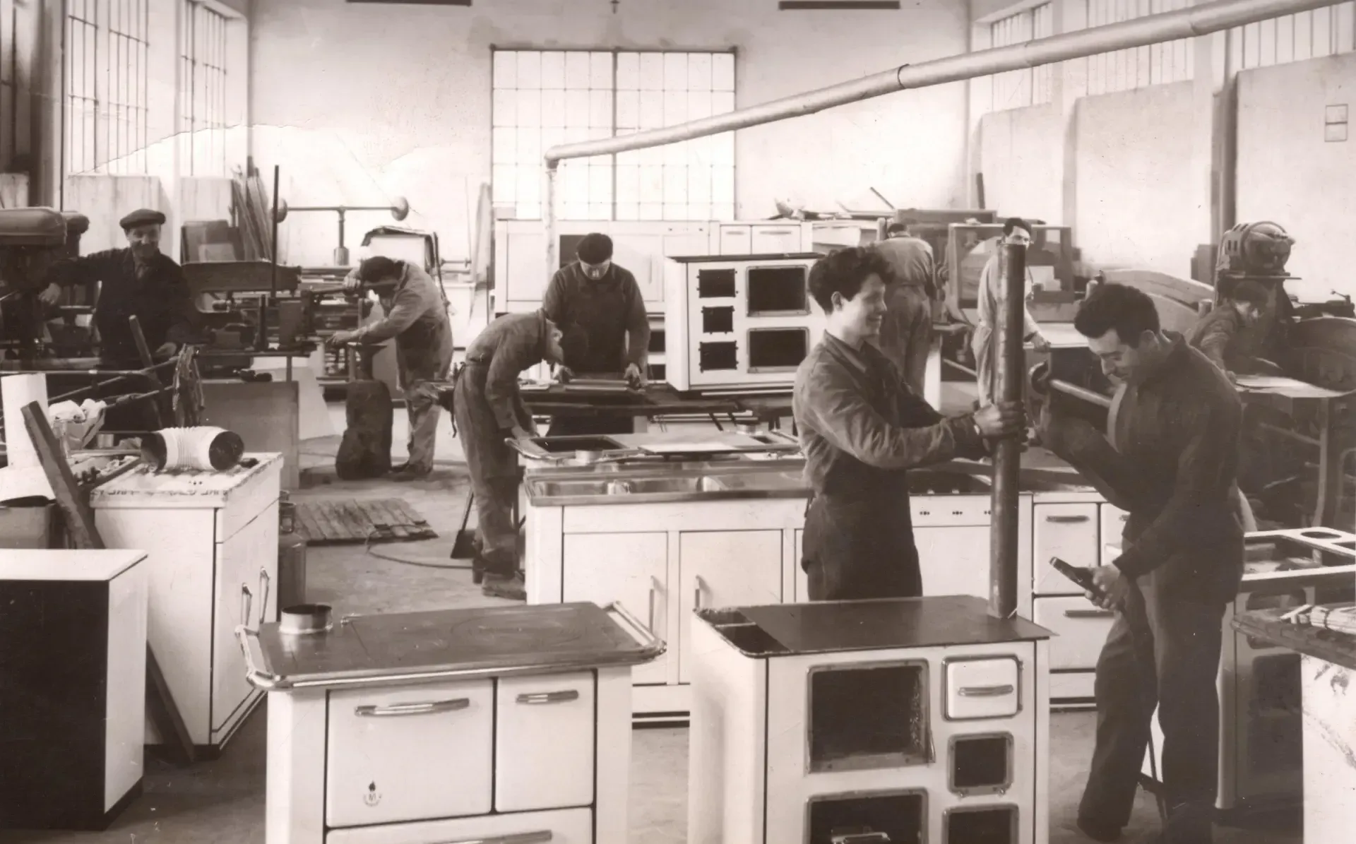 Officina con diversi uomini che costruiscono stufe. Lavori di carpenteria e assemblaggio in corso.