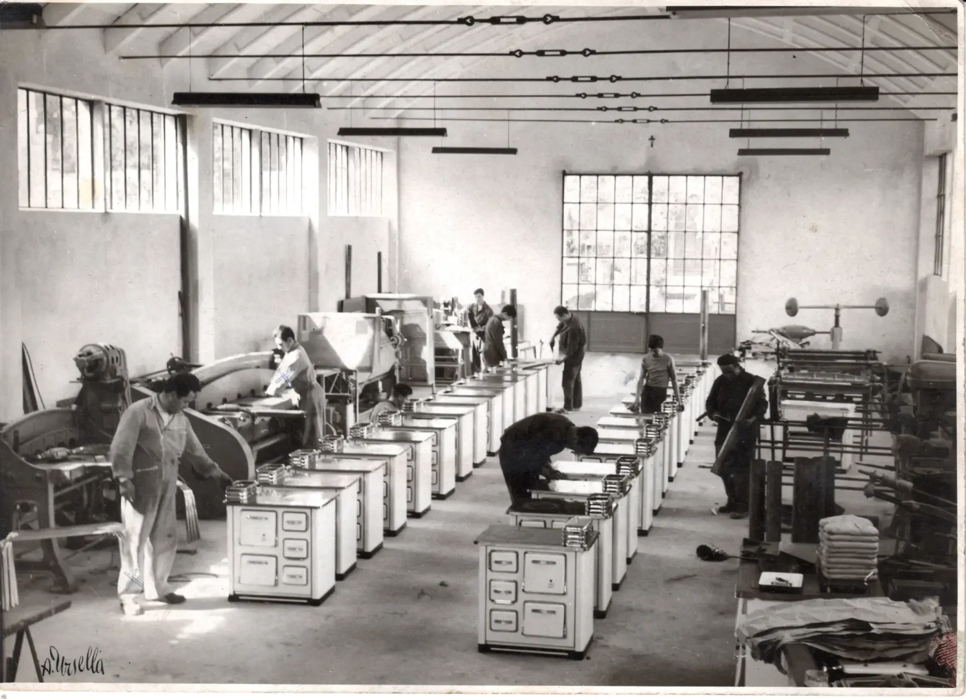 Reparto di fabbrica con operai che assemblano stufe. File di stufe, uomini al lavoro, grandi finestre.