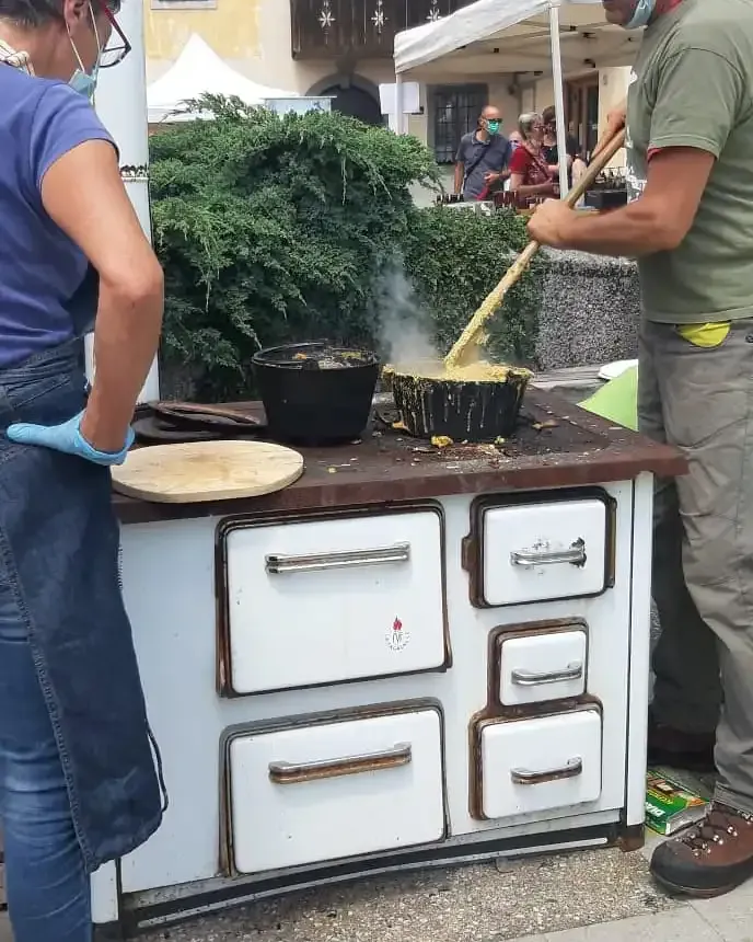 Persone che cucinano all'aperto su una stufa bianca con pentole nere. Uno mescola nella pentola, l'altro indossa un grembiule.