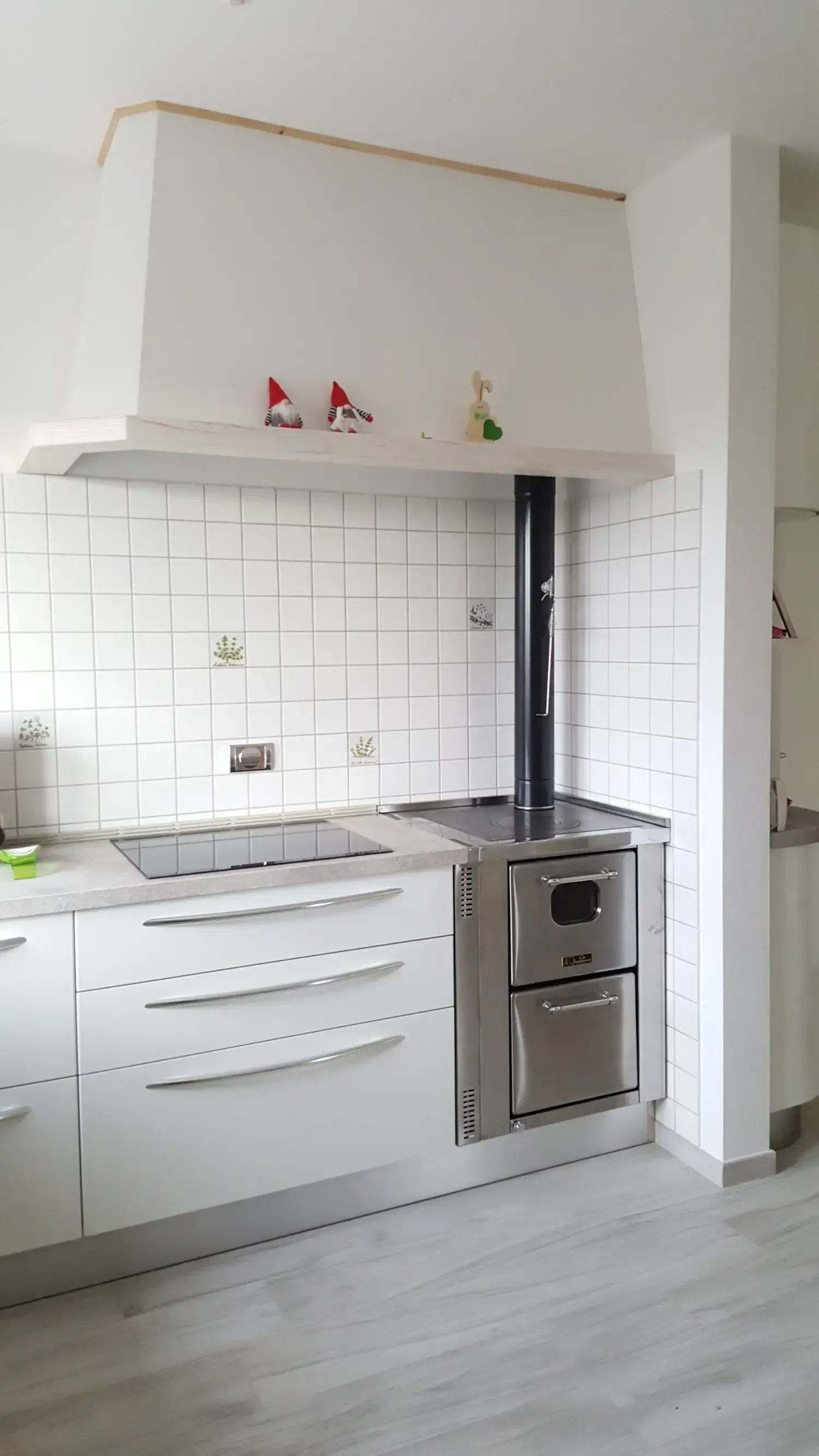 Cucina con mobili bianchi, fornelli e camino contro una parete piastrellata bianca.