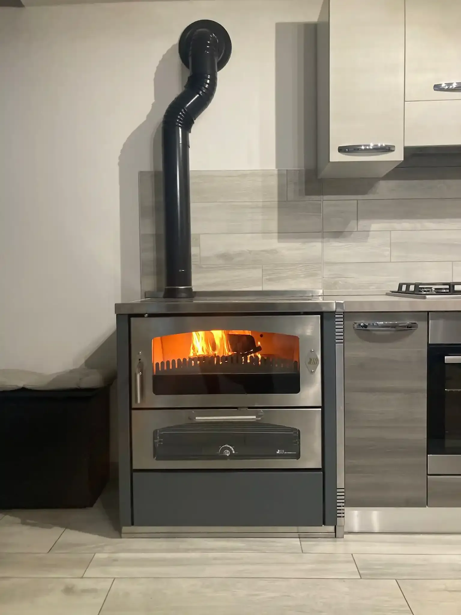 Stufa a legna con fuoco acceso in cucina; canna fumaria nera, pannelli in acciaio inossidabile e grigi.