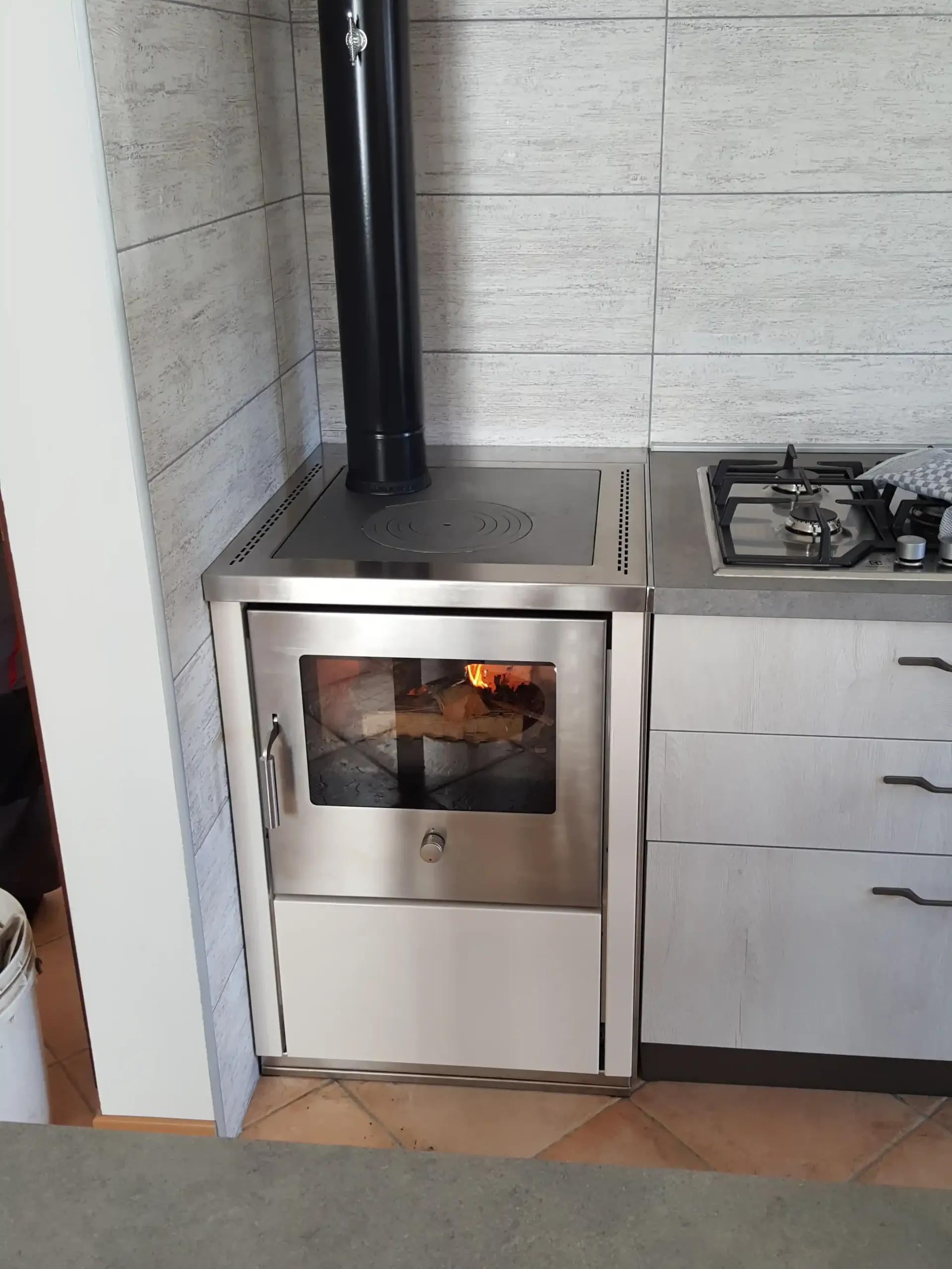 Stufa a legna in acciaio inox con fiamme visibili nello sportello del forno, accanto a un piano cottura a gas e mobili bianchi.