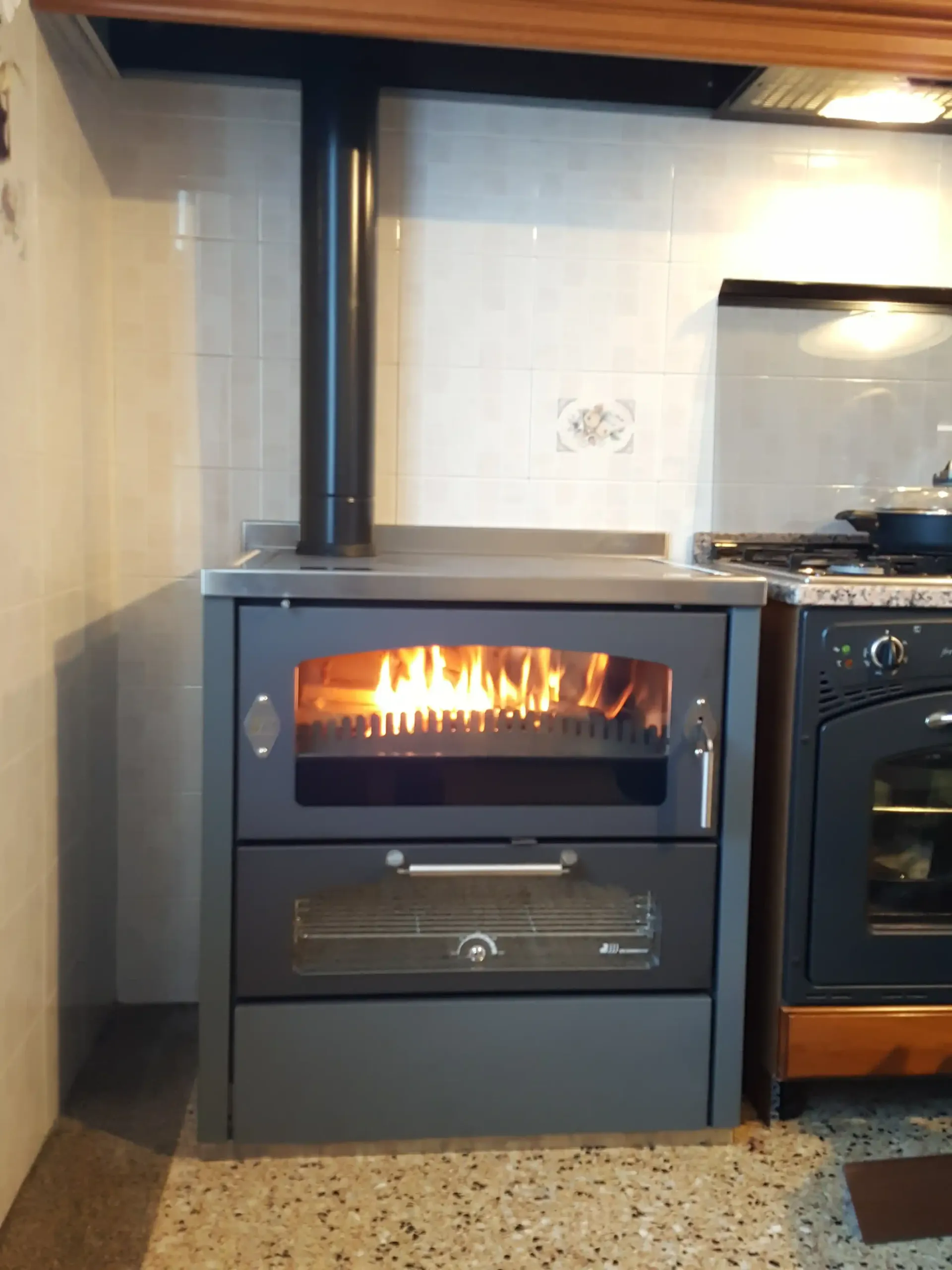 Stufa a legna con fiamme nel focolare, forno sottostante. Camino nero. Collocata in cucina.