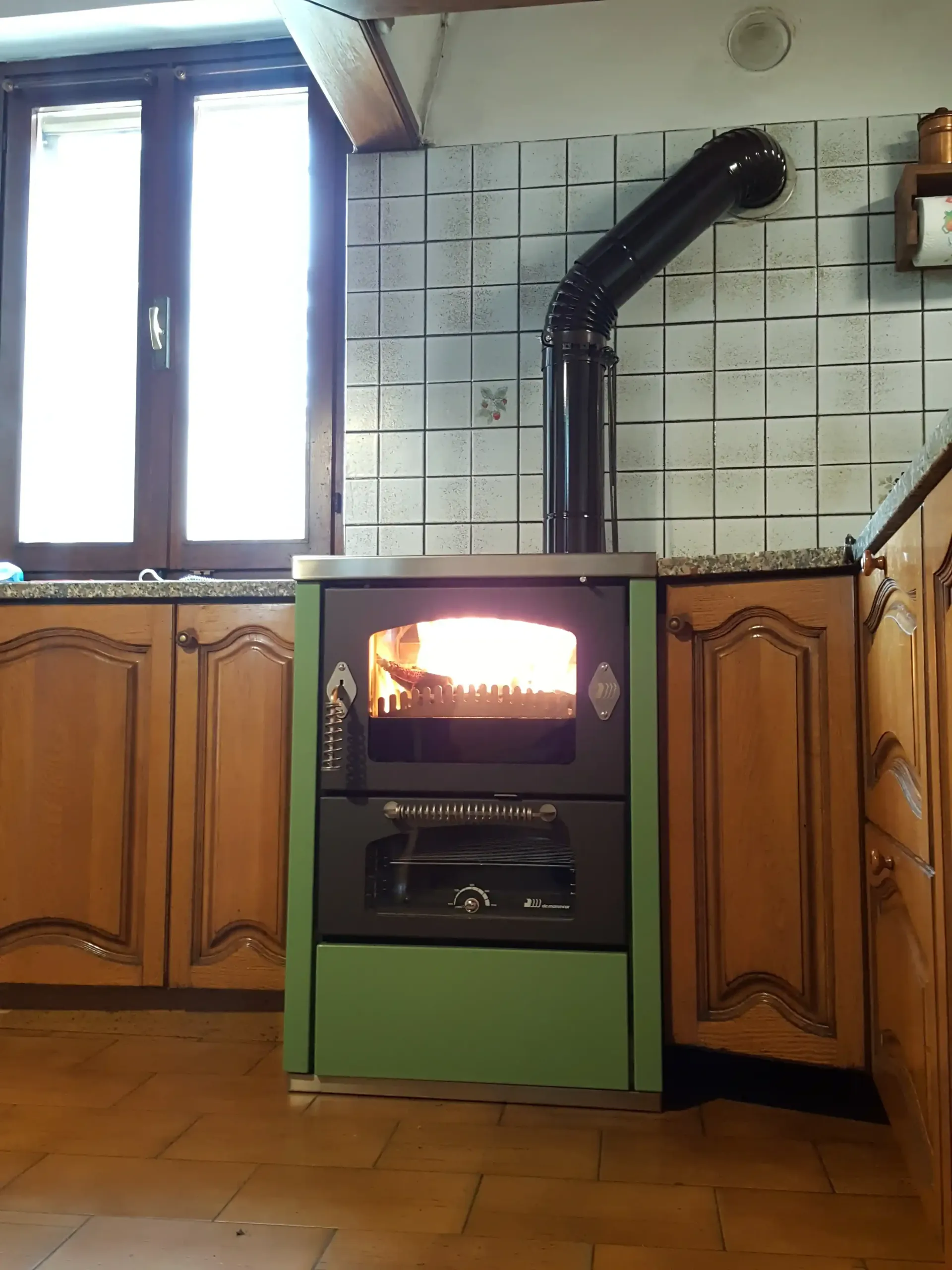 Stufa a legna verde in una cucina con camino aperto. Camino, paraschizzi piastrellato, mobili in legno e finestra.