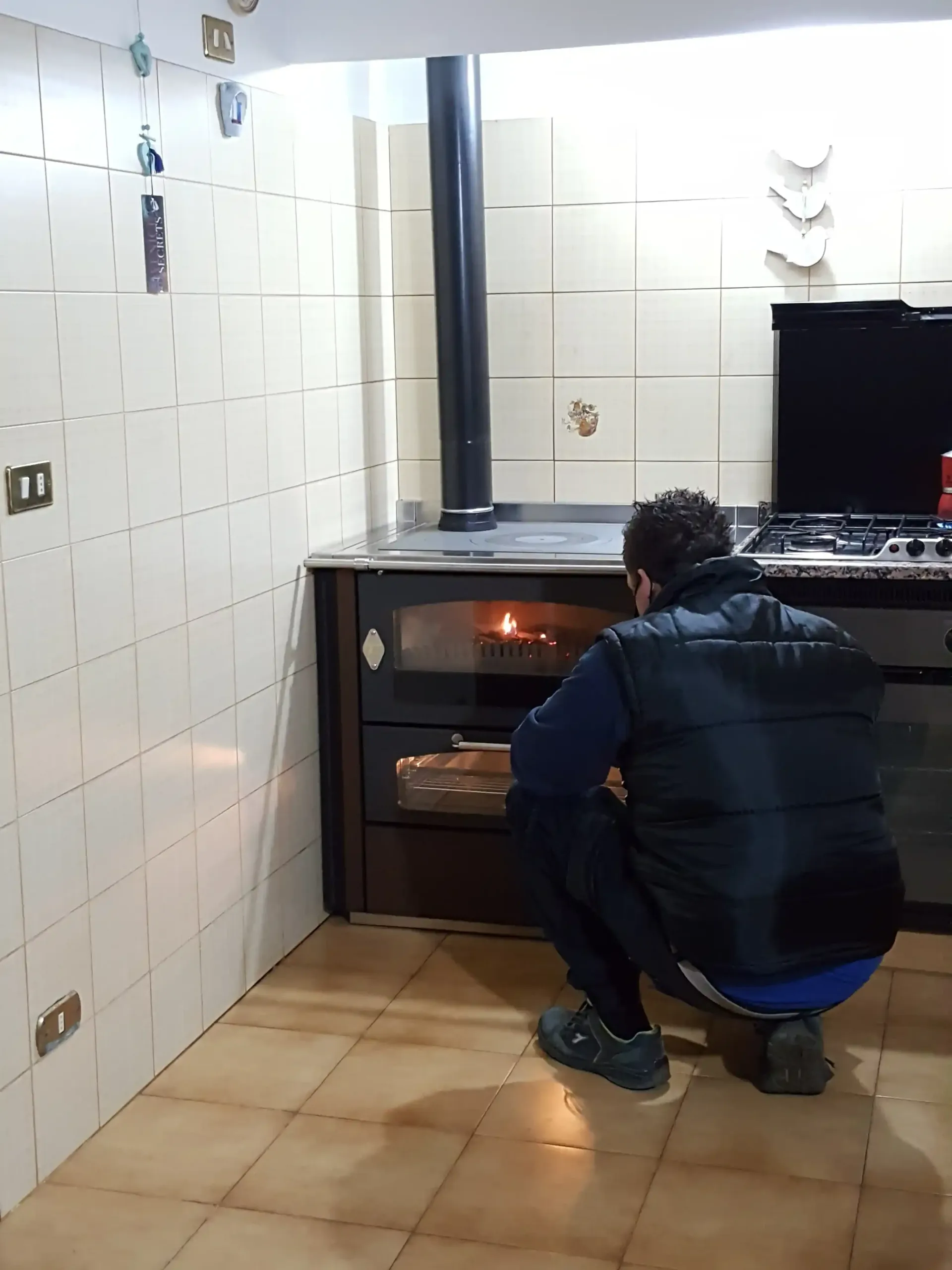 Persona accovacciata che guarda dentro un forno a legna in una cucina con pareti e pavimento piastrellati.