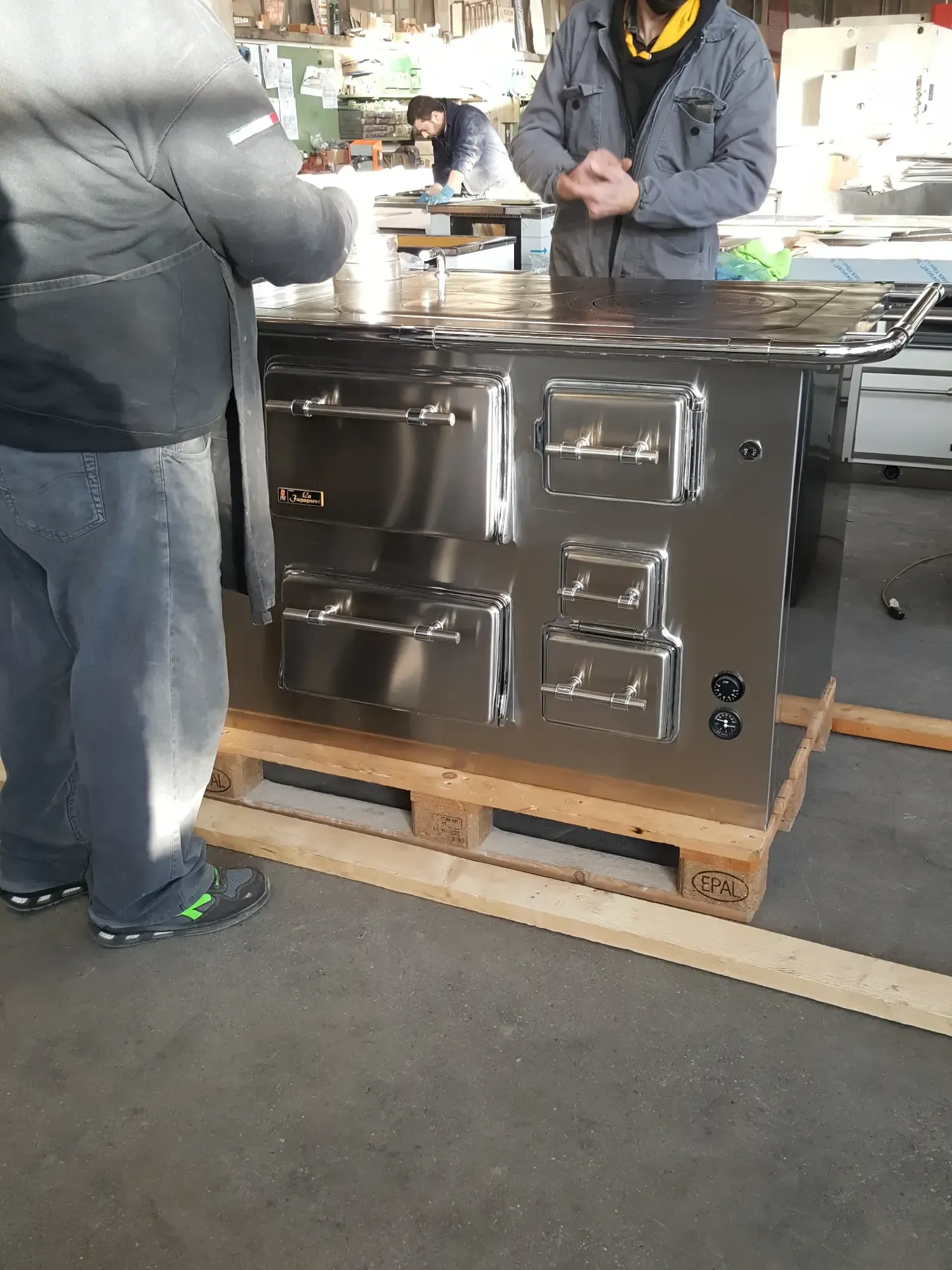 Forno commerciale in acciaio inossidabile su un pallet; due persone in piedi vicino ad esso in un magazzino.