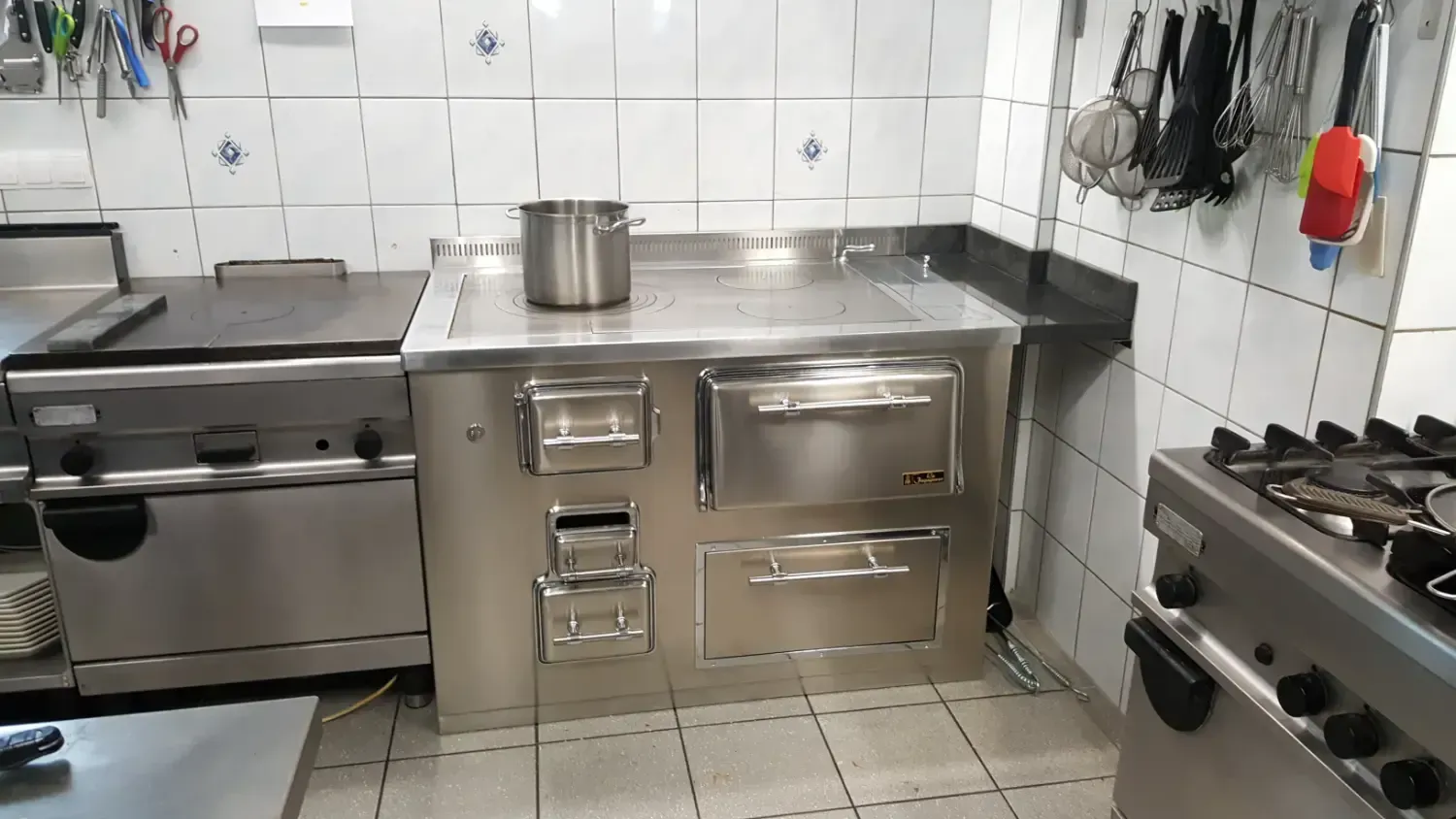 Cucina commerciale con piano cottura, forno e pentole in acciaio inox; pareti piastrellate bianche.