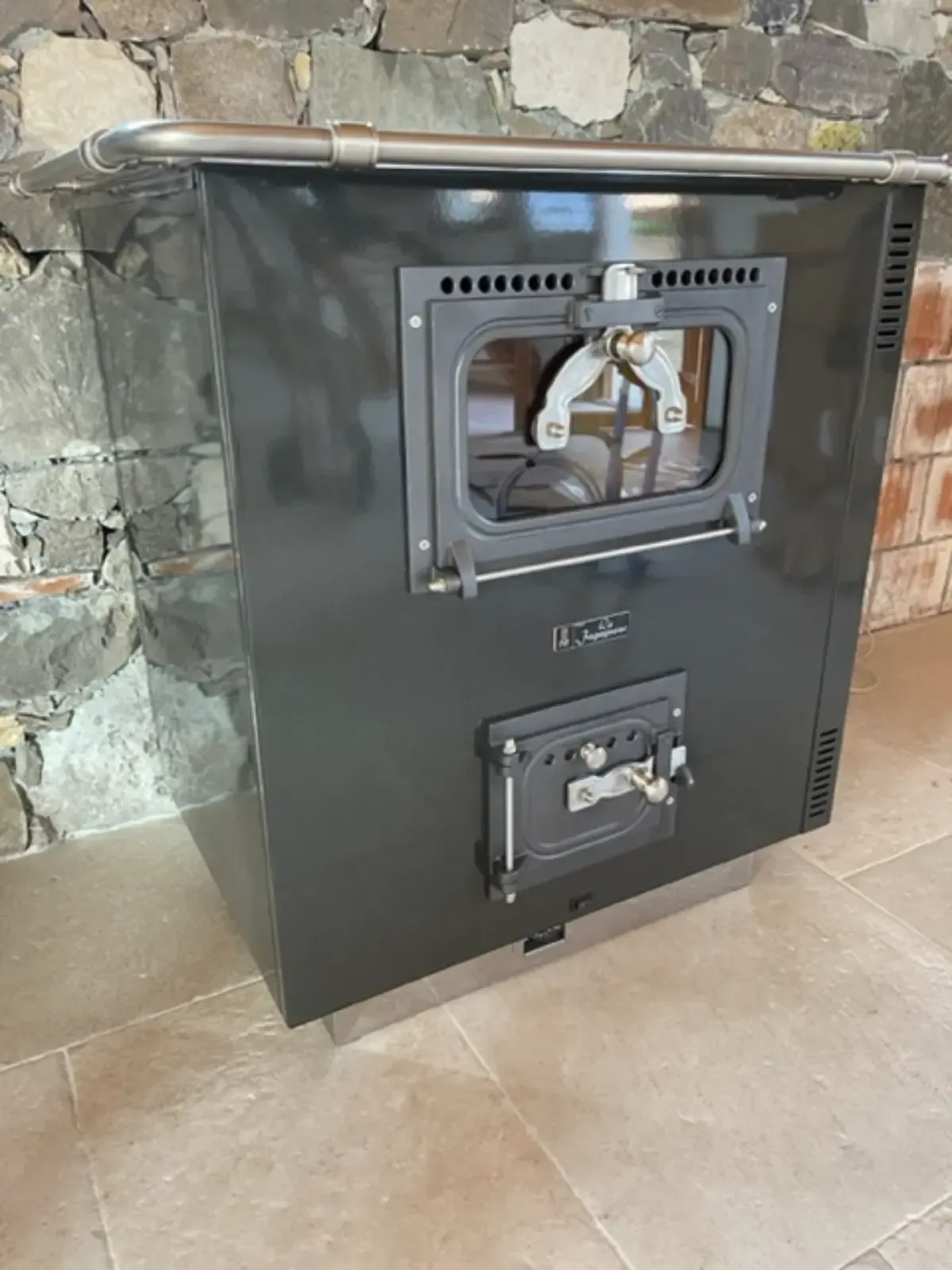 Stufa a legna nera con sportello del forno in vetro e sportello di accesso inferiore, appoggiata a pareti in pietra e mattoni.