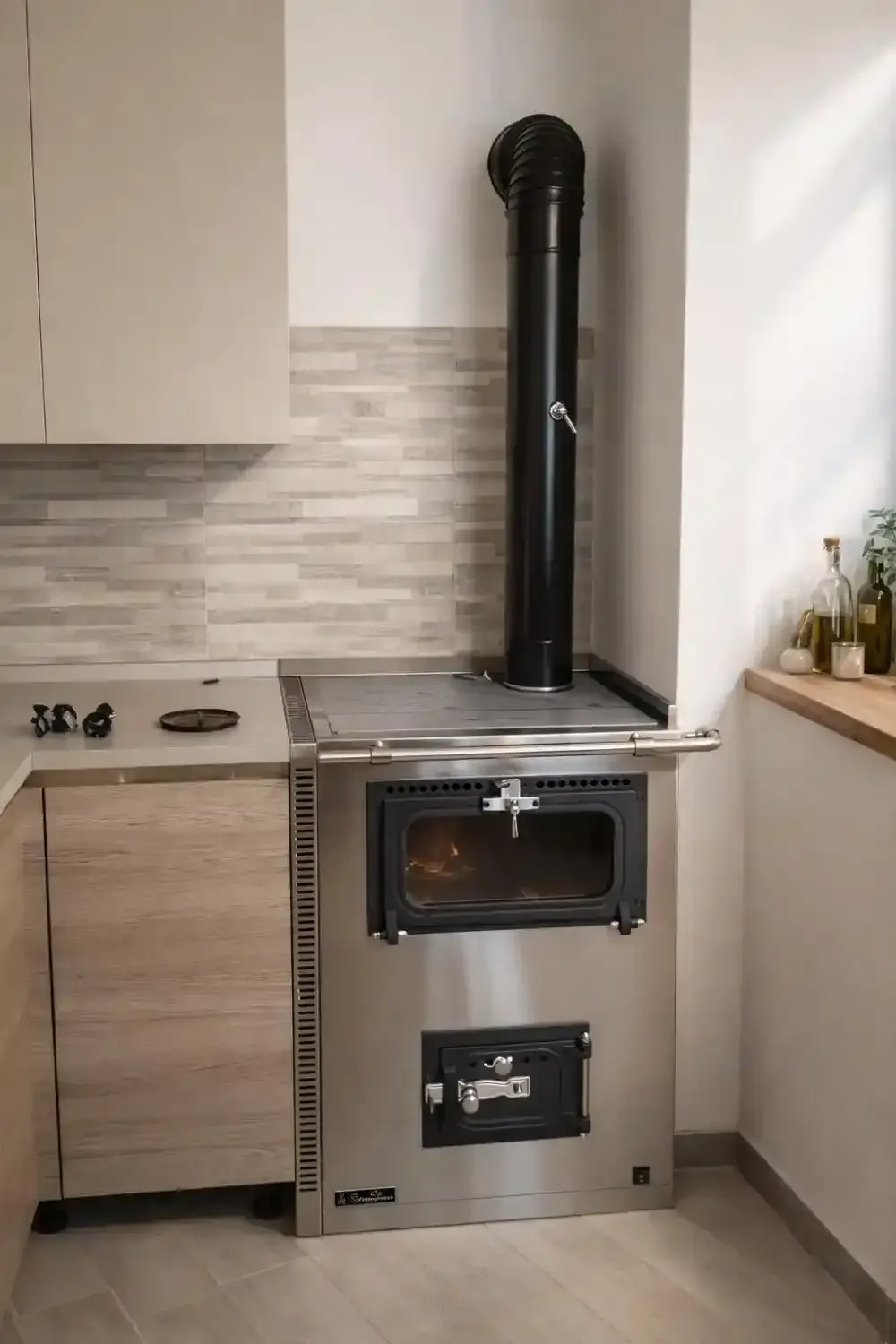 Stufa a legna in acciaio inossidabile con camino nero in una cucina accanto a un bancone e dei mobili.