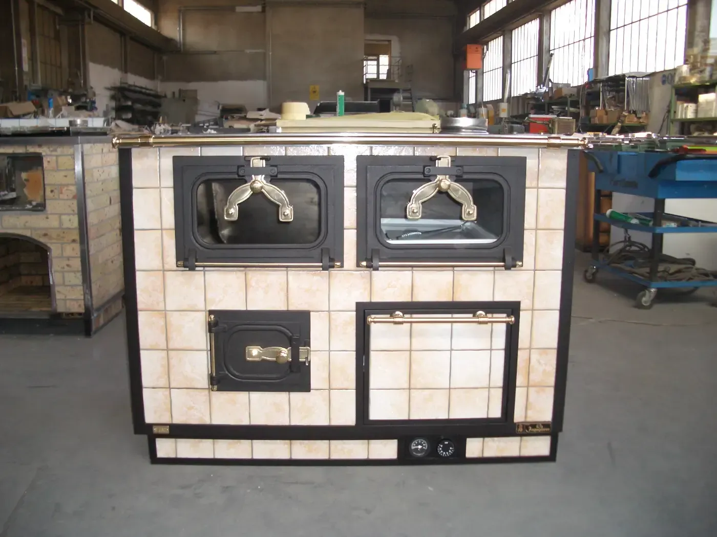 Forno in maiolica con due sportelli e un piccolo sportello inferiore. Dettagli dorati. In un ambiente industriale.