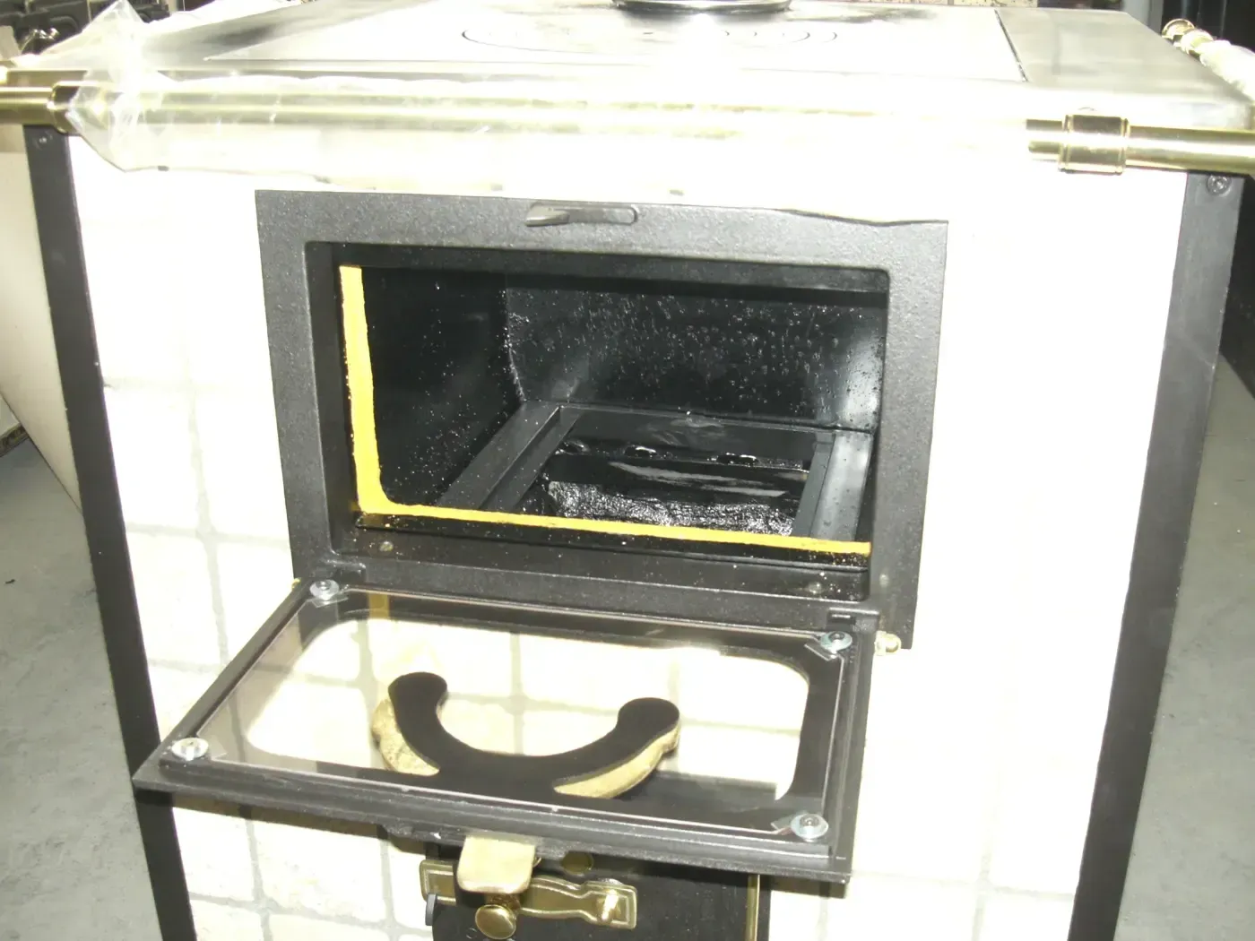 Stufa a legna aperta, esterno piastrellato bianco, interno nero, guarnizione gialla intorno alla porta, porta aperta con maniglia.
