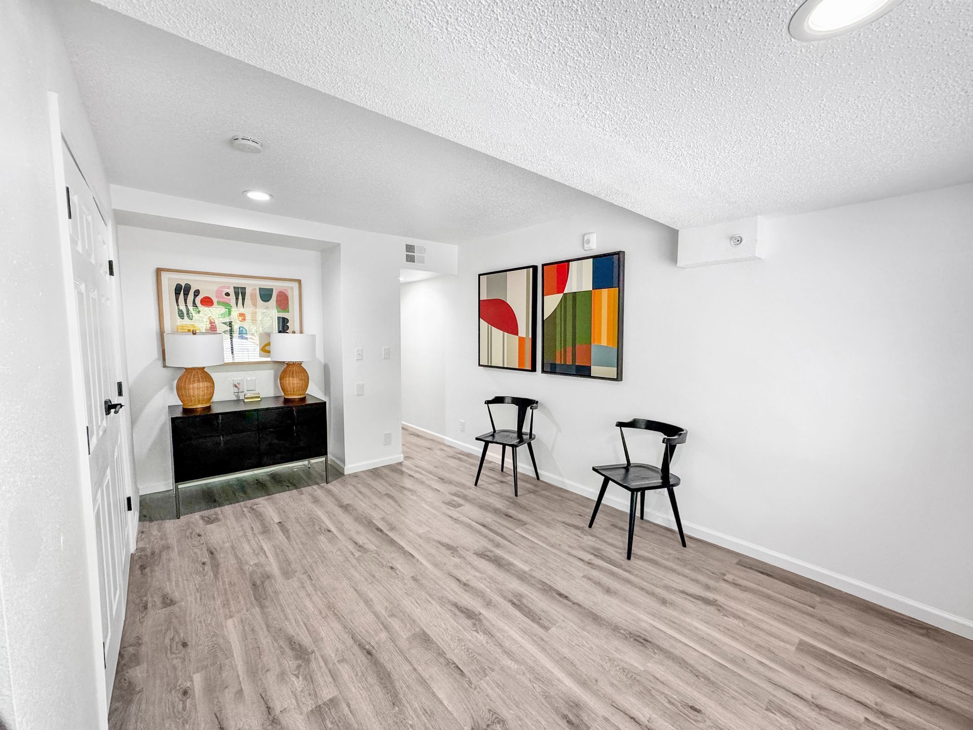 Bright minimalist room with wood floors, abstract wall art, and two stools by a black console table.
