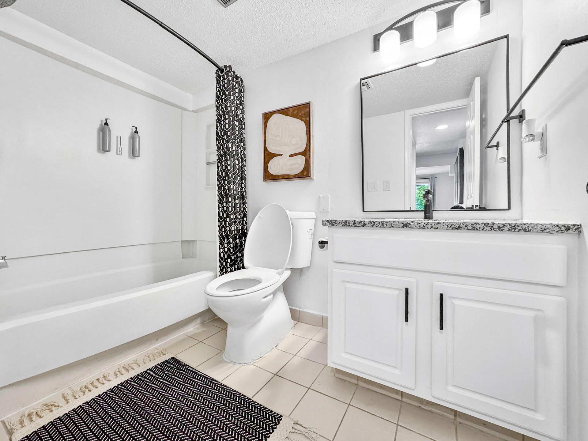Bright white bathroom with tub, toilet, vanity, mirror, and black-and-white rug.
