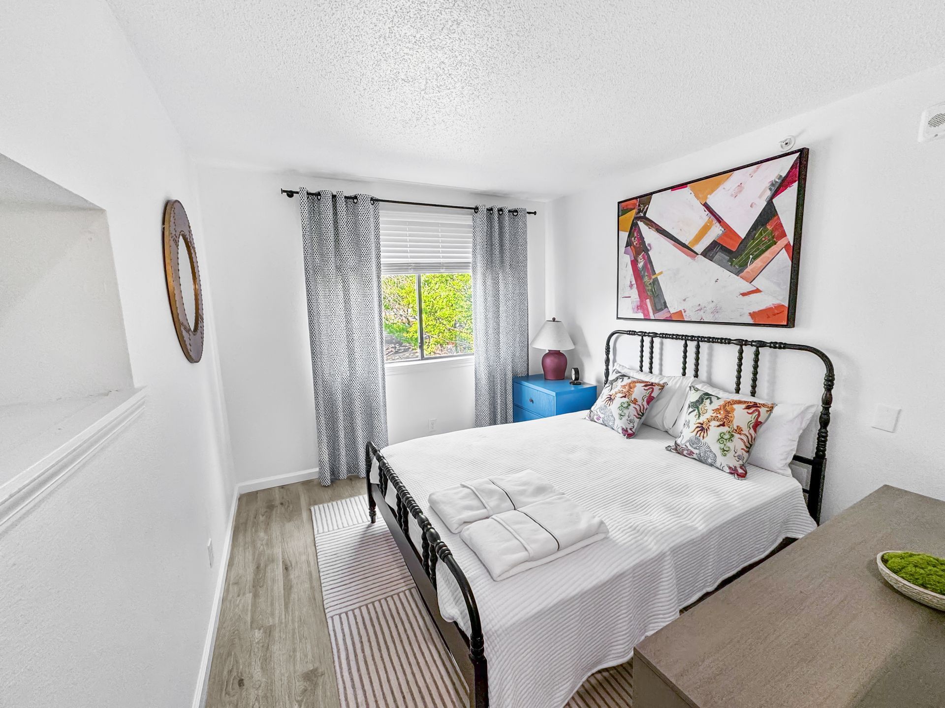 Bright bedroom with a white bed, patterned curtains, colorful wall art, and a blue dresser by the window