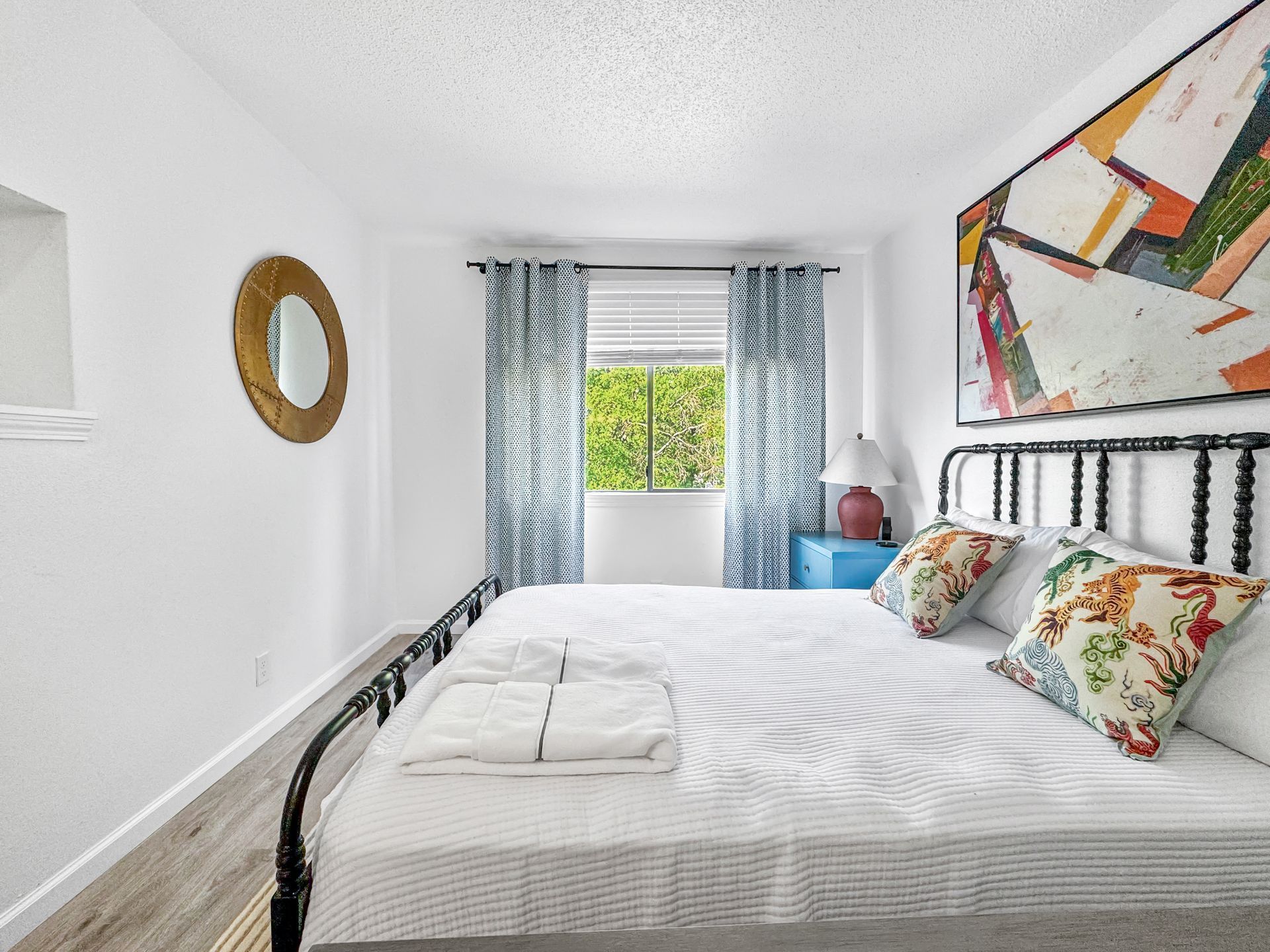 Bright bedroom with white bedspread, patterned curtains, and colorful wall art by a window