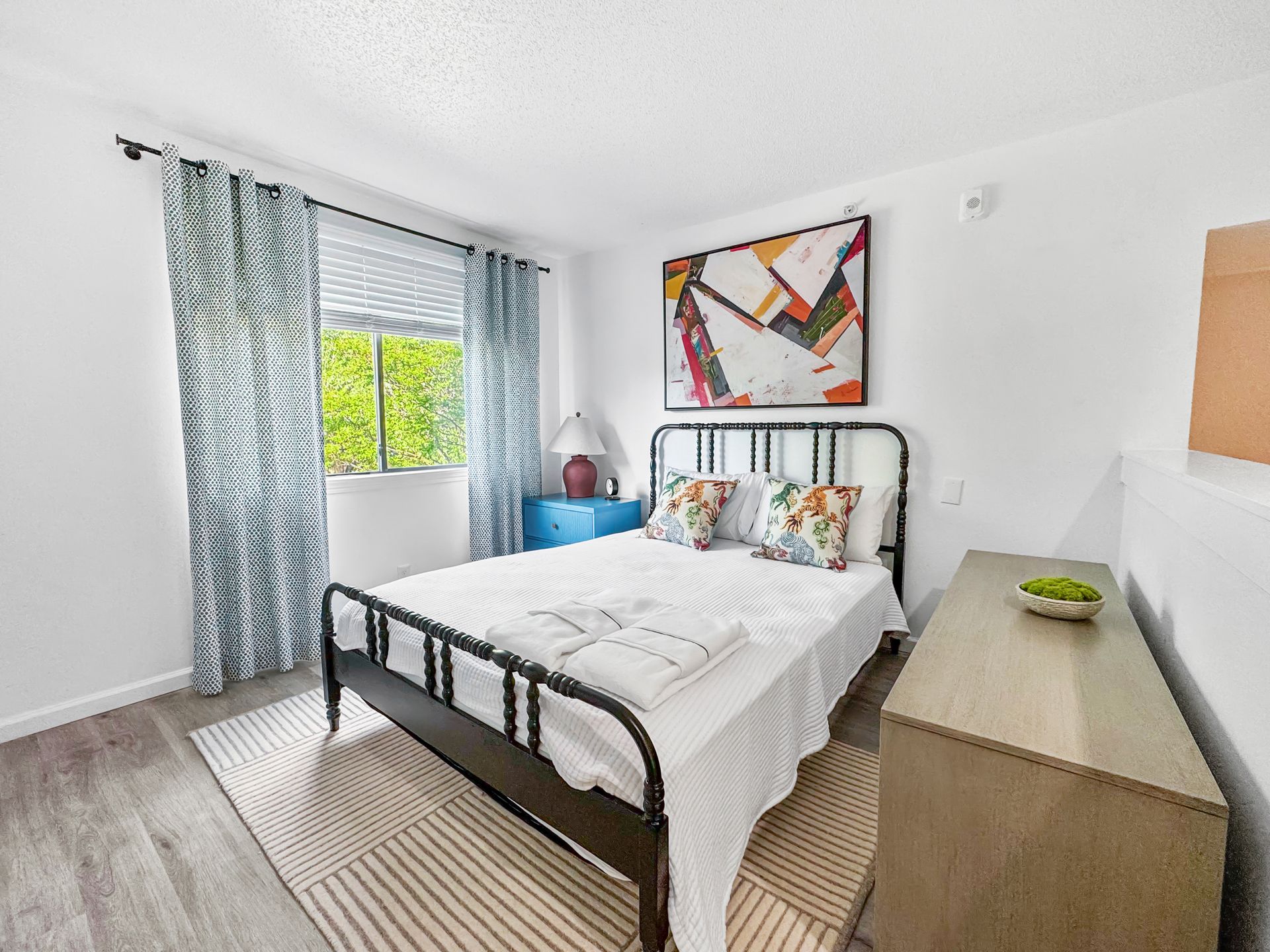 Bright bedroom with a black bed frame, blue curtains, colorful wall art, and a wooden dresser