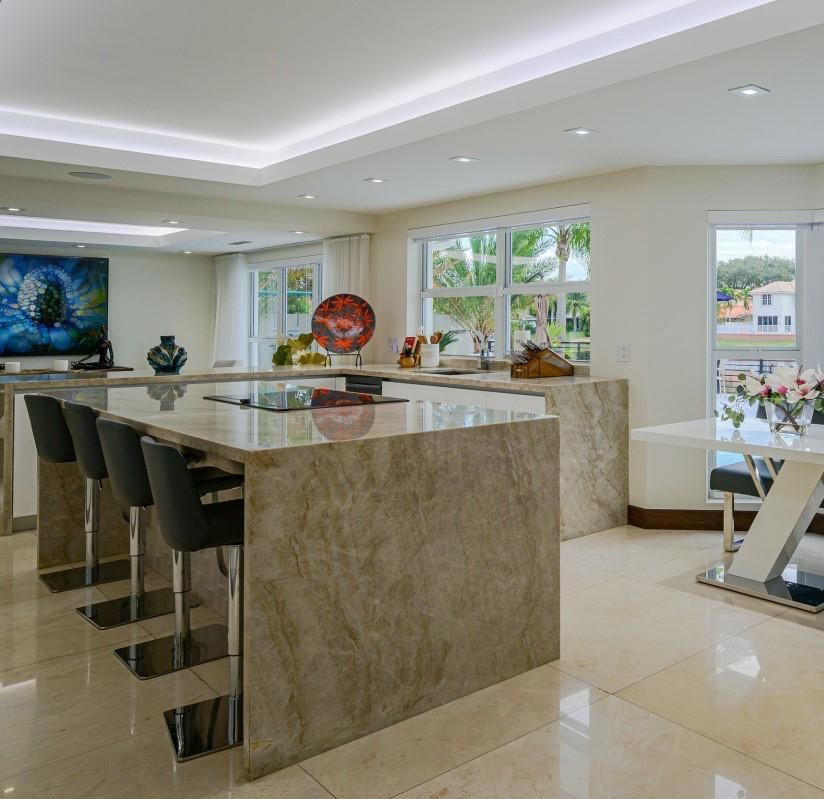 Modern kitchen with marble island, bar stools, glossy tile floor, and large windows overlooking a bright room.