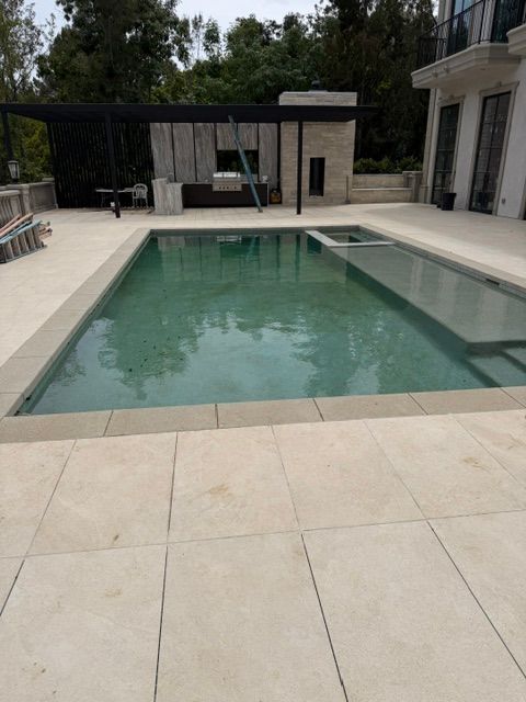 Rectangular backyard swimming pool with pale stone patio and covered seating area beside a modern house