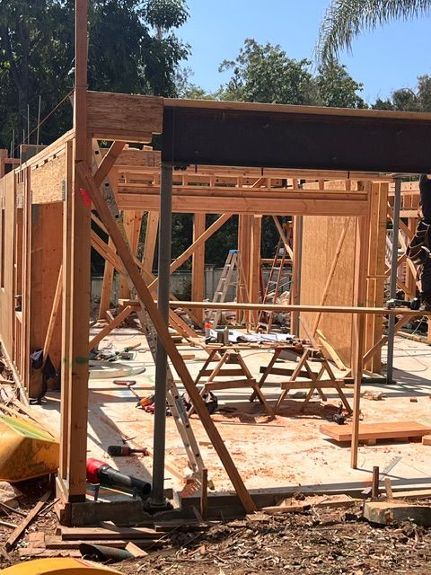 Wood-framed house under construction with scaffolding, beams, and a temporary roof in a sunny outdoor site