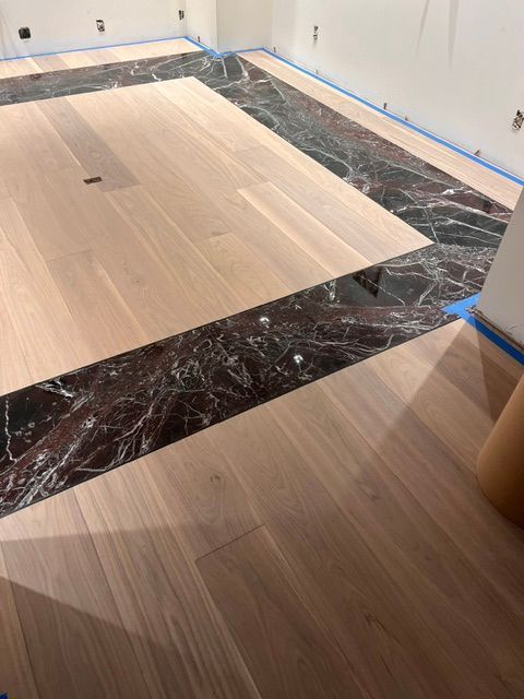Partially installed light wood flooring with blue tape around the room and a dark marble border strip